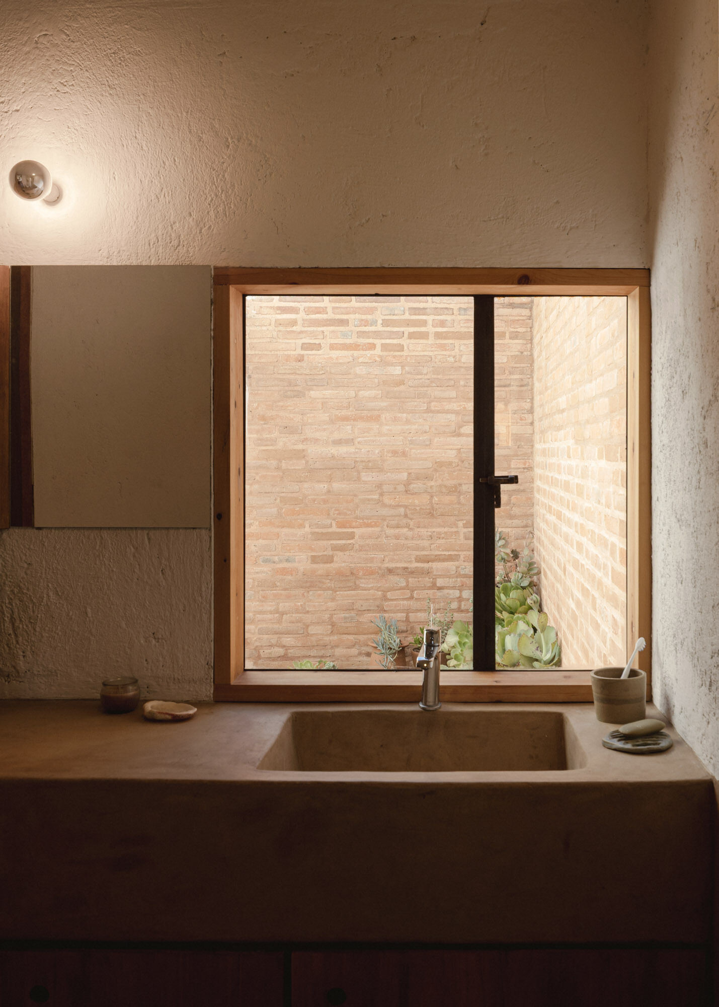Casa Amona’s bathroom pairs a built-in basin and cypress storage with a window overlooking exposed brick and a small planted space.