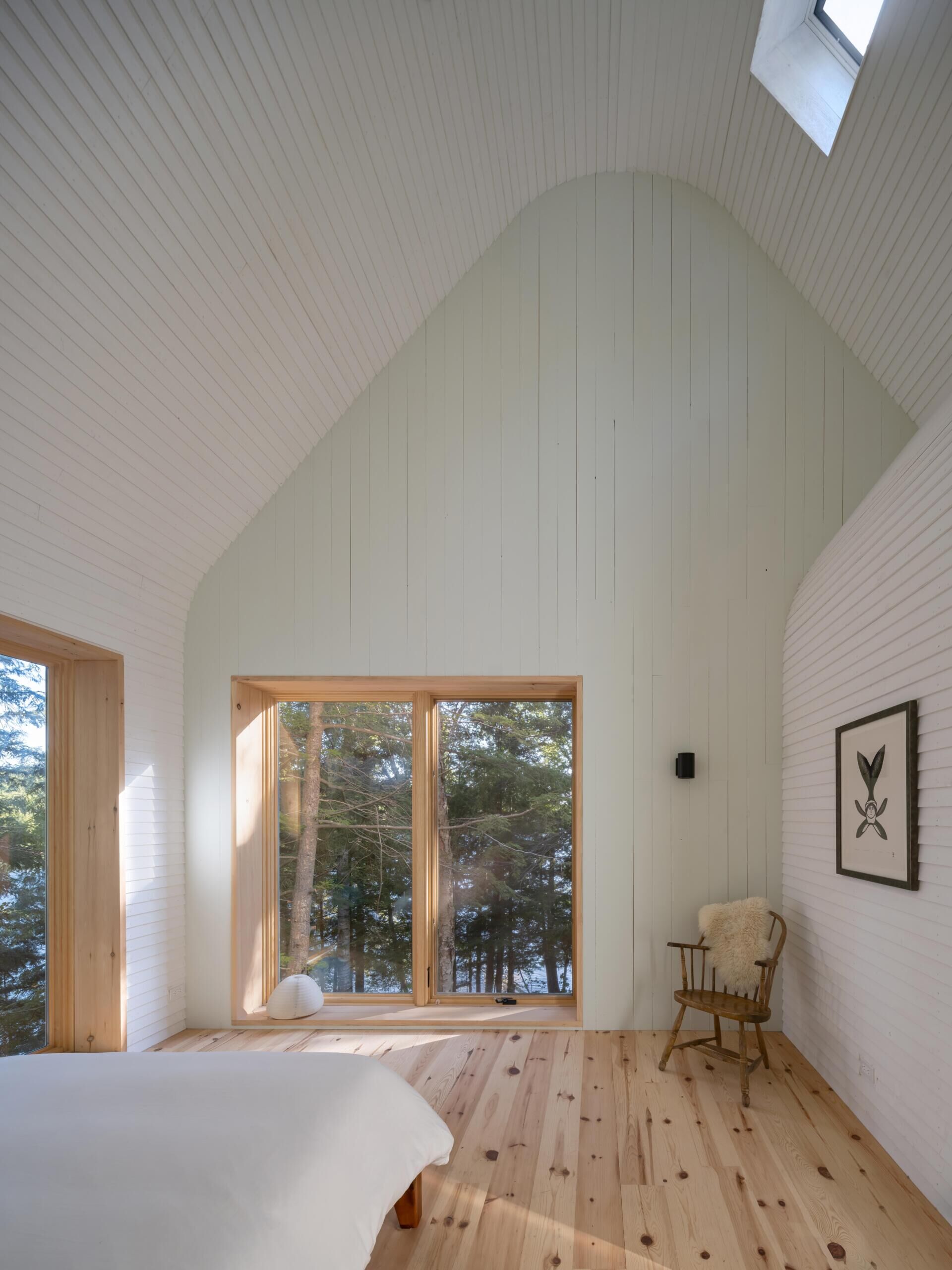 A minimalist cottage bedroom with vaulted ceilings, white timber walls, pale green gable details, warm wood floors, and large windows overlooking the forest landscape.