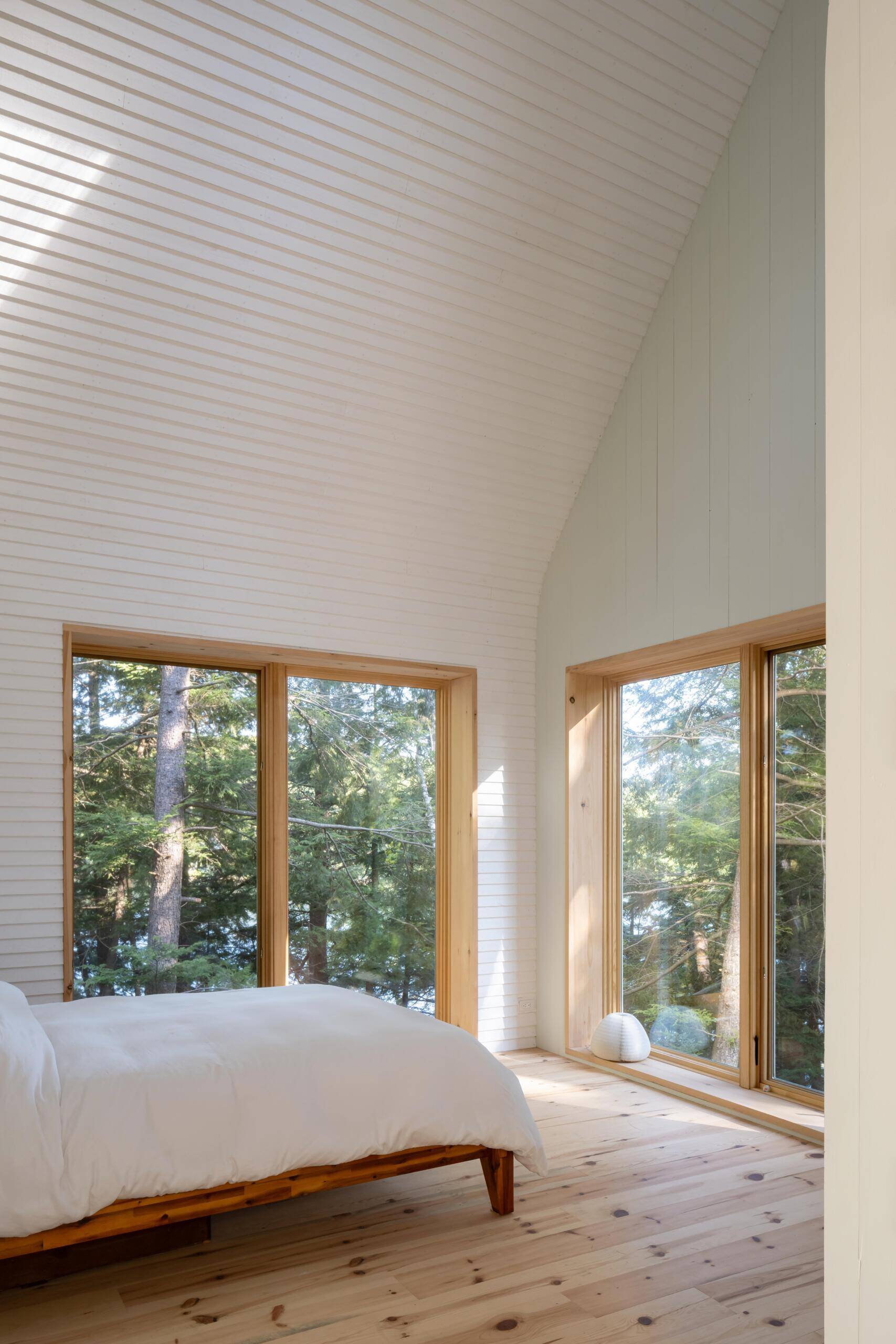 A minimalist cottage bedroom with vaulted ceilings, white timber walls, pale green gable details, warm wood floors, and large windows overlooking the forest landscape.