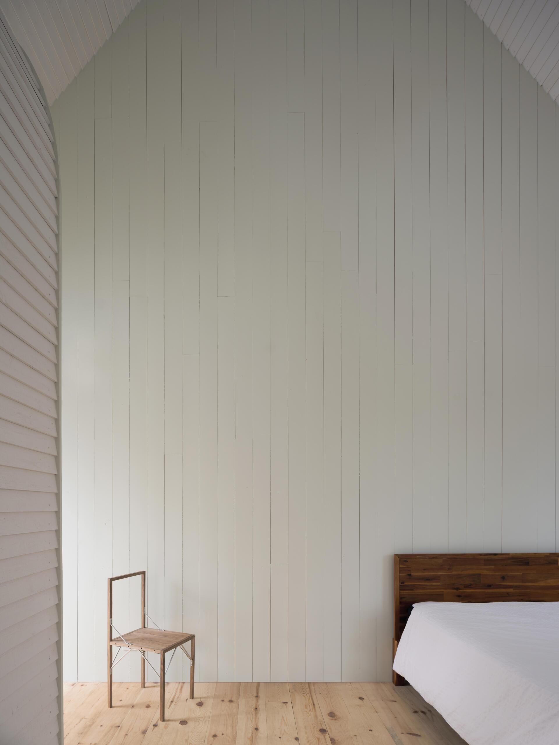 A minimalist cottage bedroom with vaulted ceilings, white timber walls, pale green gable details, warm wood floors, and large windows overlooking the forest landscape.
