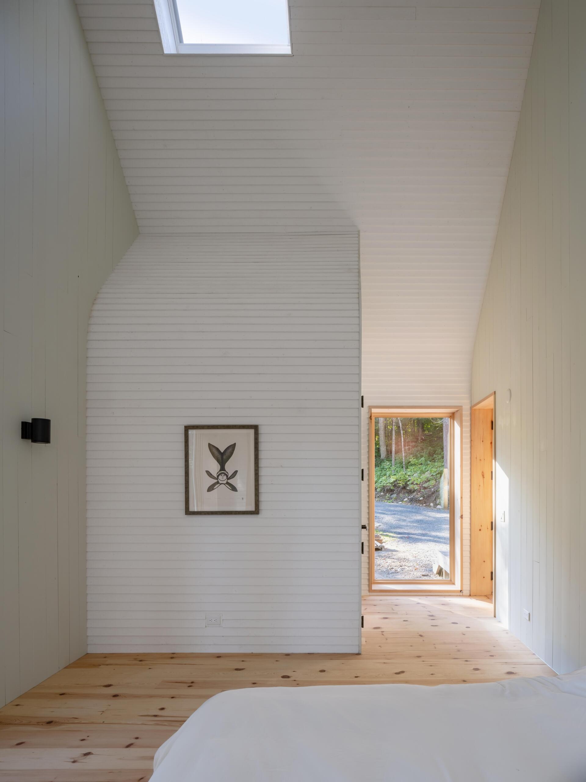 A minimalist cottage bedroom with vaulted ceilings, white timber walls, pale green gable details, warm wood floors, and large windows overlooking the forest landscape.
