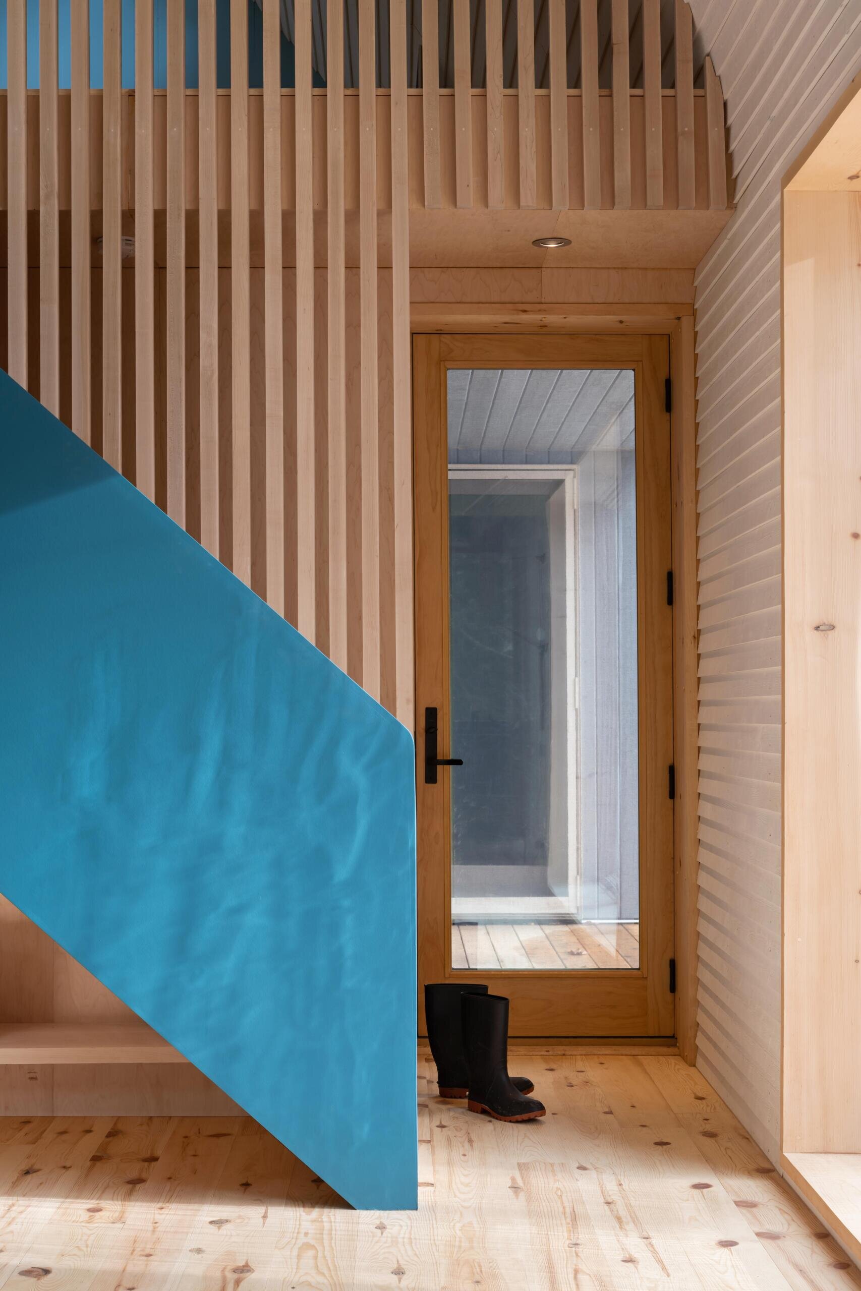 A modern cottage entryway featuring white pine walls, warm timber details, a maple slat screen, and a bold blue stair wall that adds colour to the minimalist interior.