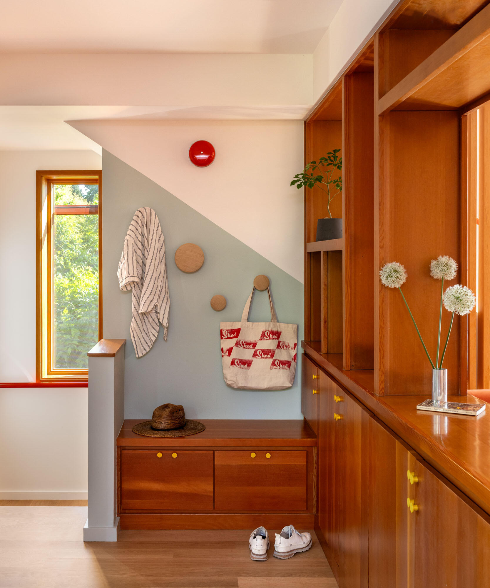See how SHED transformed a wall of formal dark cabinetry into warm built-in storage with open display niches, yellow knobs, a bench with drawers and circular hooks for coats and bags.