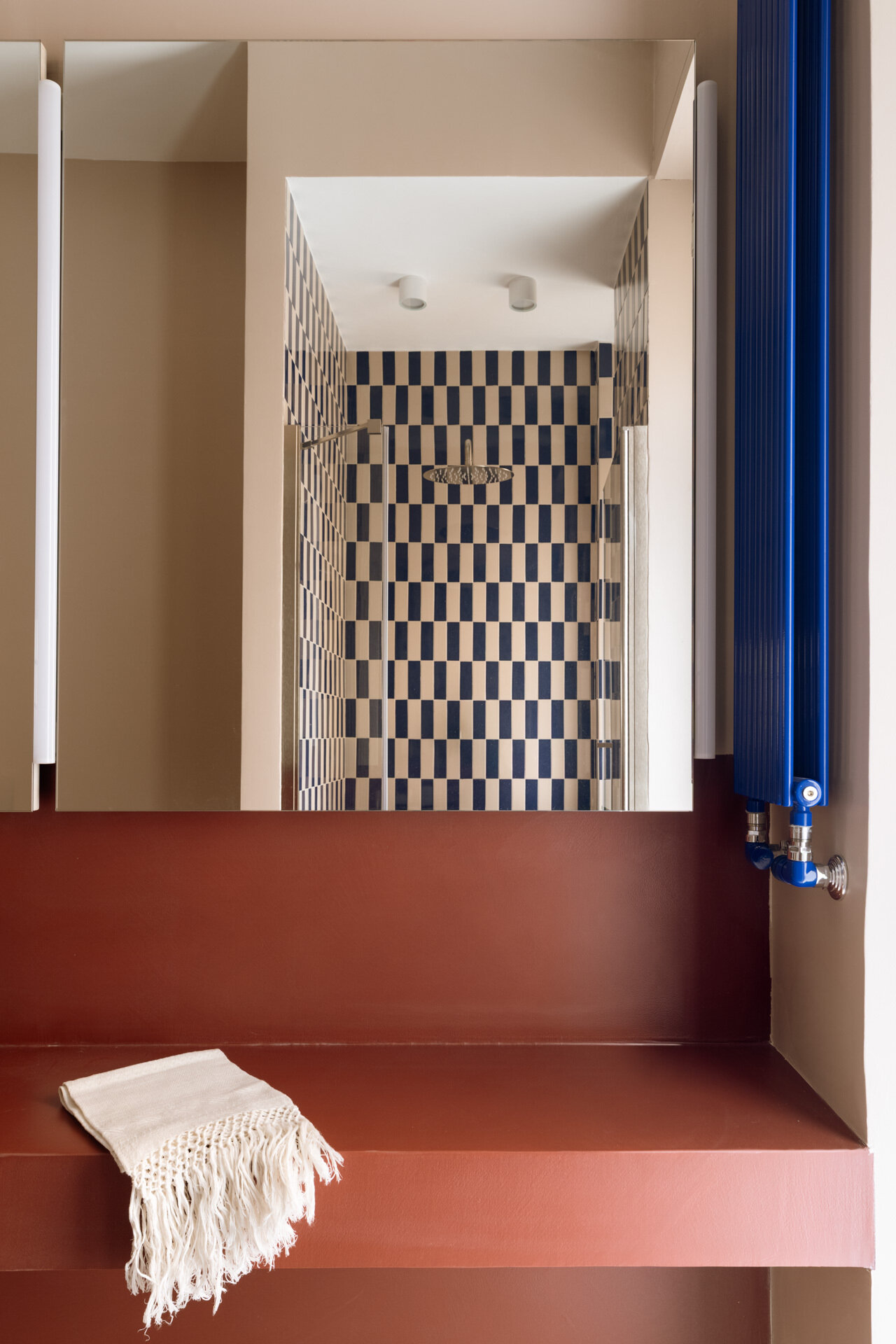 A colorful contemporary bathroom features a floating vanity, white vessel sink, large illuminated mirrors, cobalt-blue radiator, and a walk-in shower with graphic blue-and-white rectangular tiles.