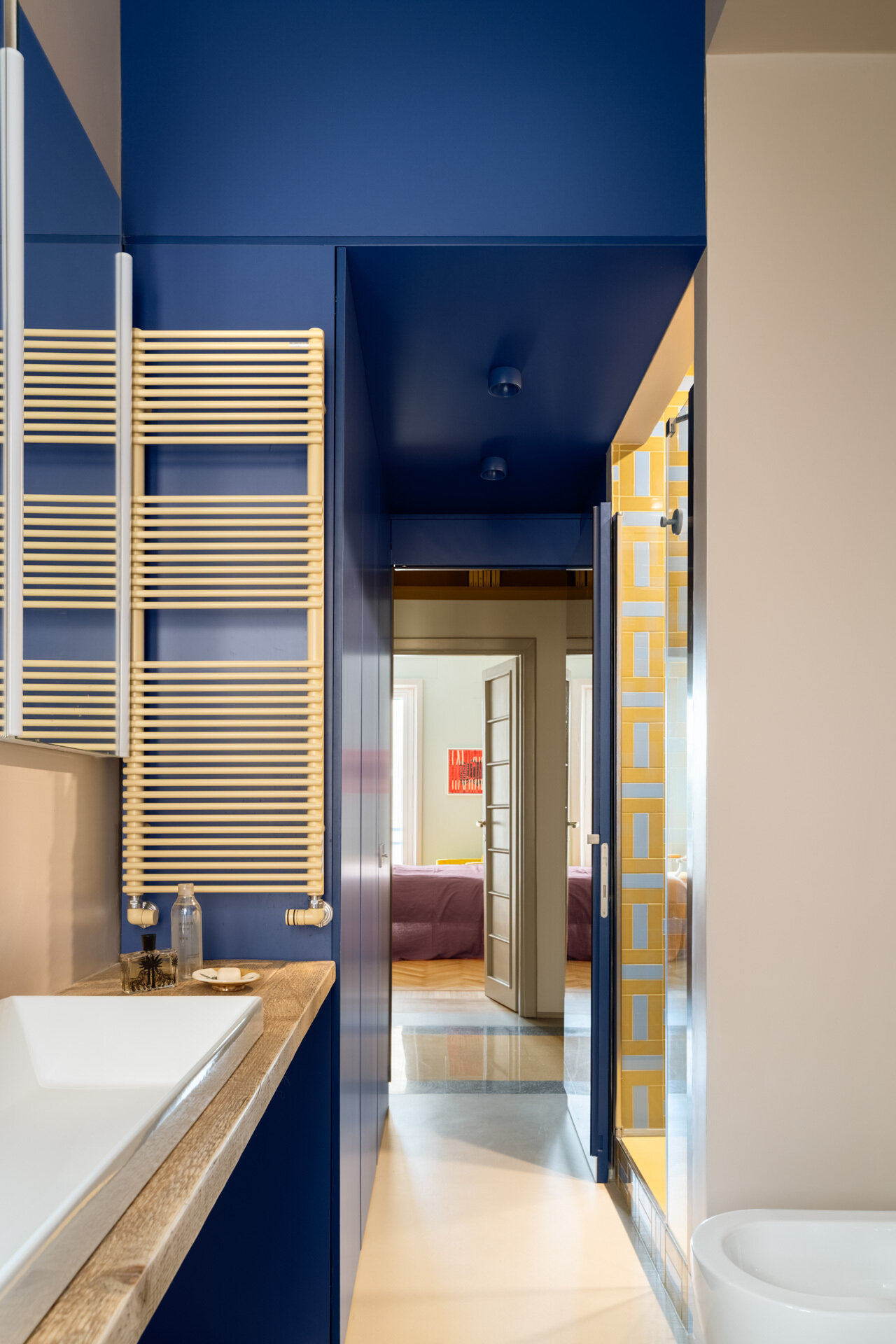 A colorful bathroom combines deep cobalt-blue walls, a long vanity countertop, a yellow towel radiator, and a walk-in shower covered in geometric yellow and pale-blue rectangular tiles.