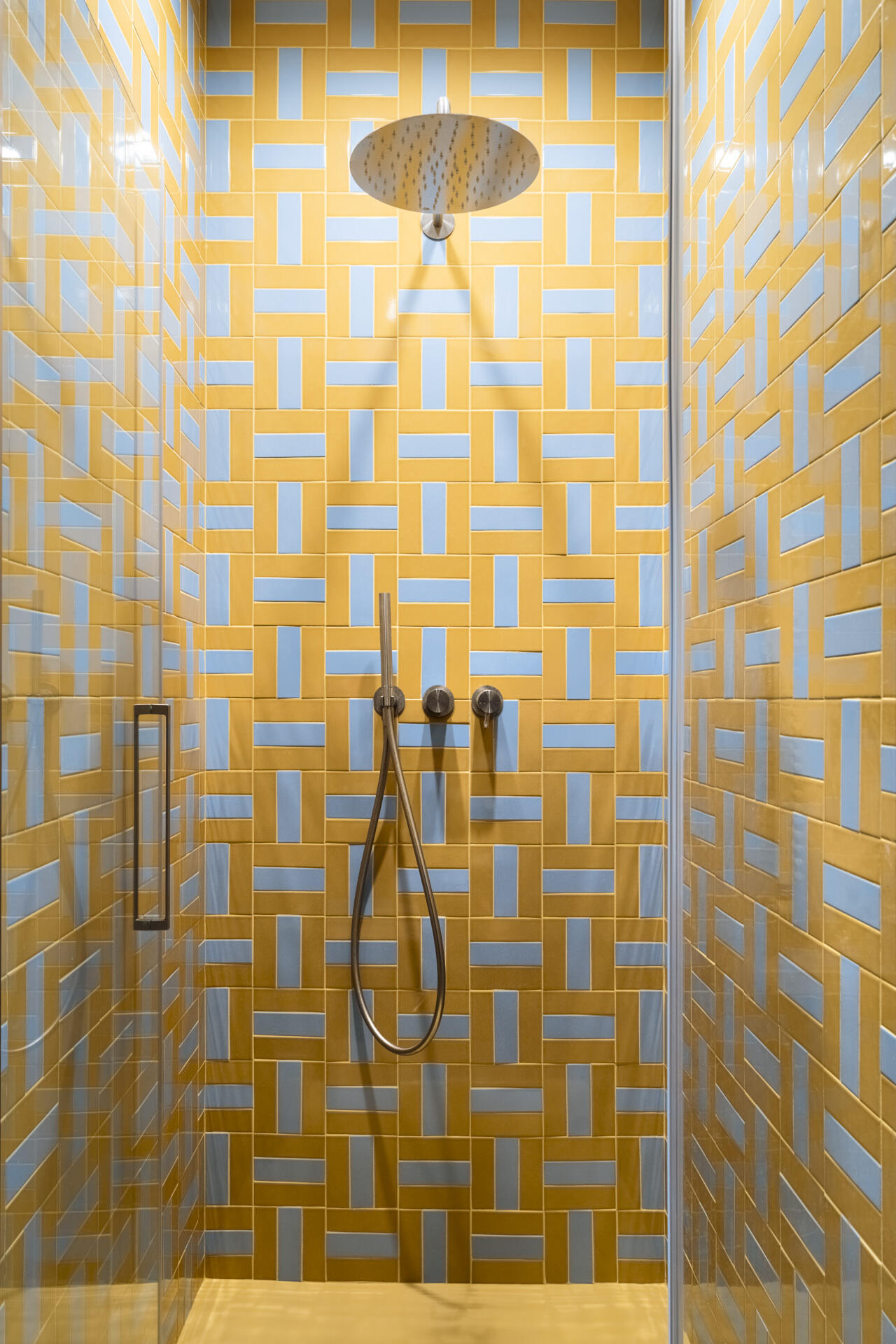 A colorful bathroom combines deep cobalt-blue walls, a long vanity countertop, a yellow towel radiator, and a walk-in shower covered in geometric yellow and pale-blue rectangular tiles.
