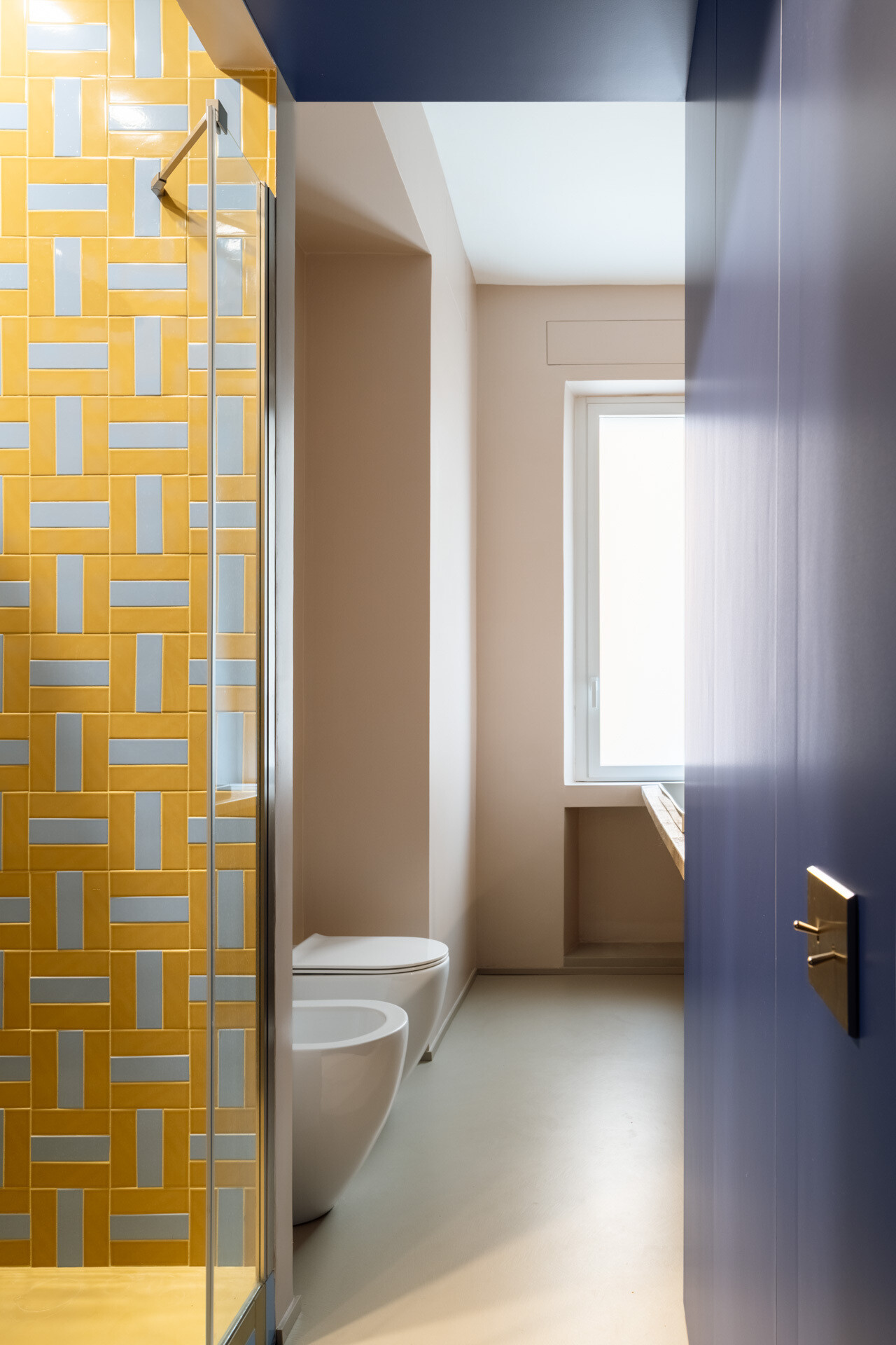 A colorful bathroom combines deep cobalt-blue walls, a long vanity countertop, a yellow towel radiator, and a walk-in shower covered in geometric yellow and pale-blue rectangular tiles.