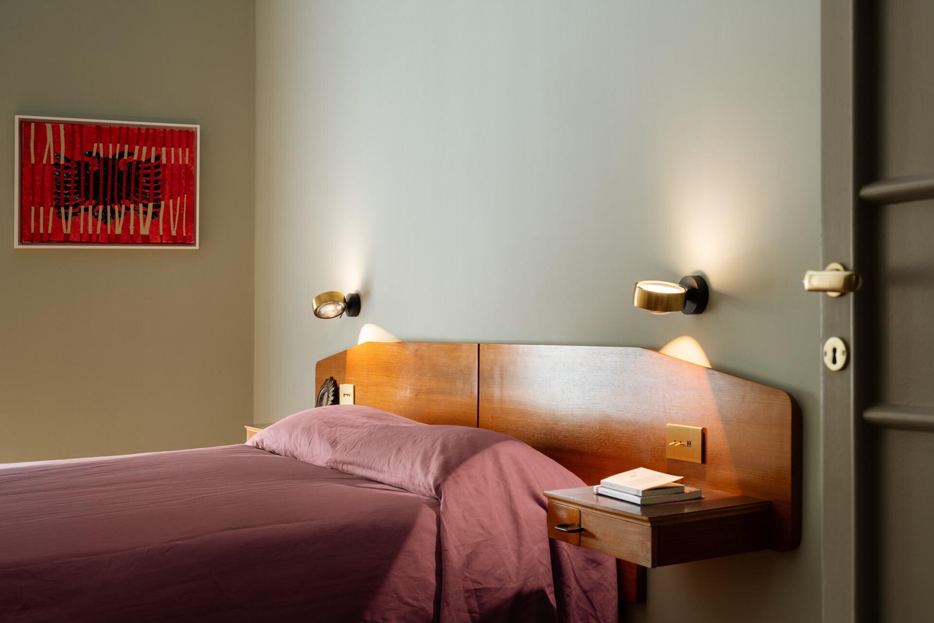 A sage green primary bedroom in Rome combines a vintage teak headboard with brass details, integrated bedside drawers, Occhio reading lights, mauve bedding, a yellow armchair, and original Italian herringbone parquet flooring.