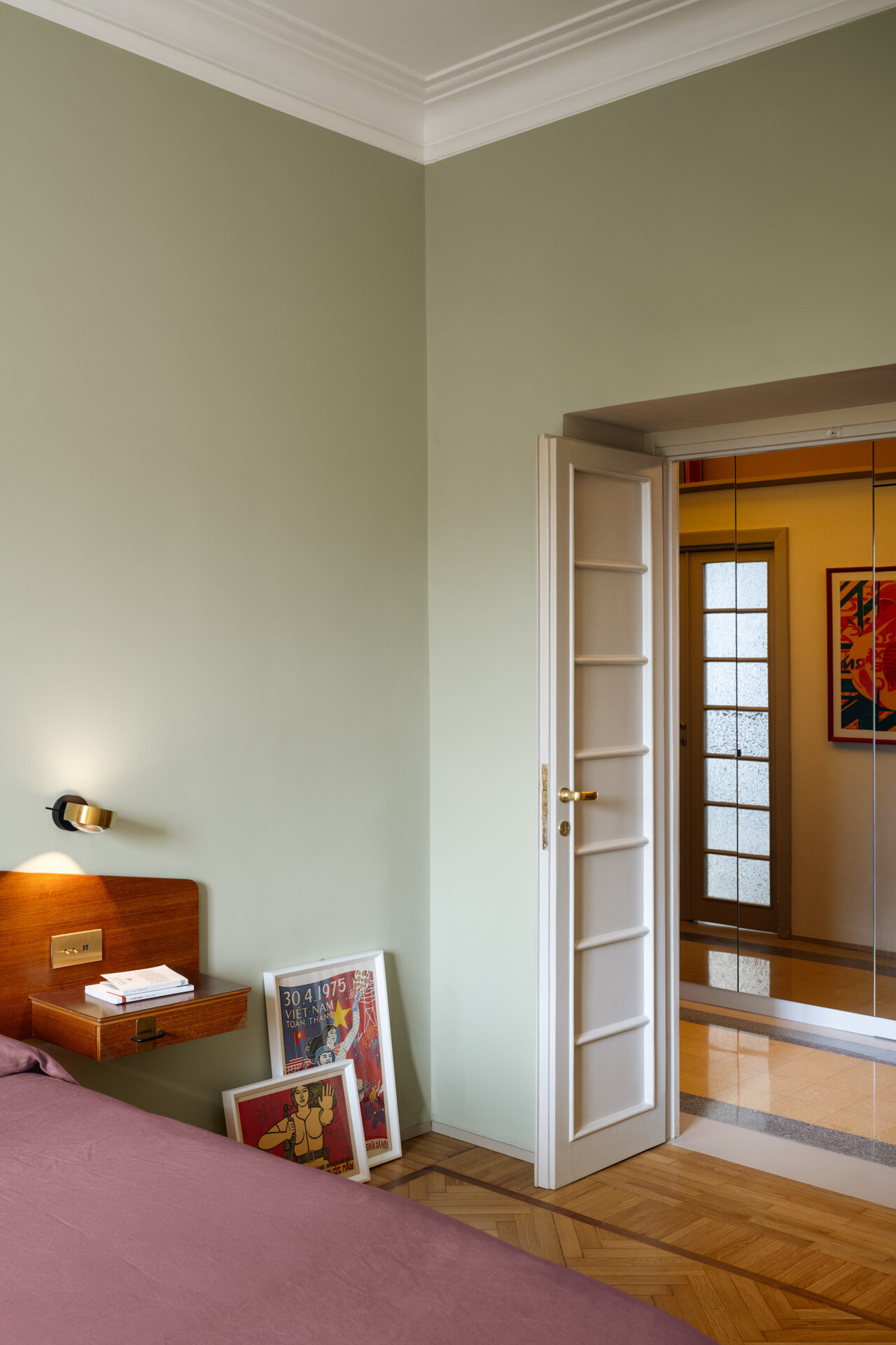 A sage green primary bedroom in Rome combines a vintage teak headboard with brass details, integrated bedside drawers, Occhio reading lights, mauve bedding, a yellow armchair, and original Italian herringbone parquet flooring.