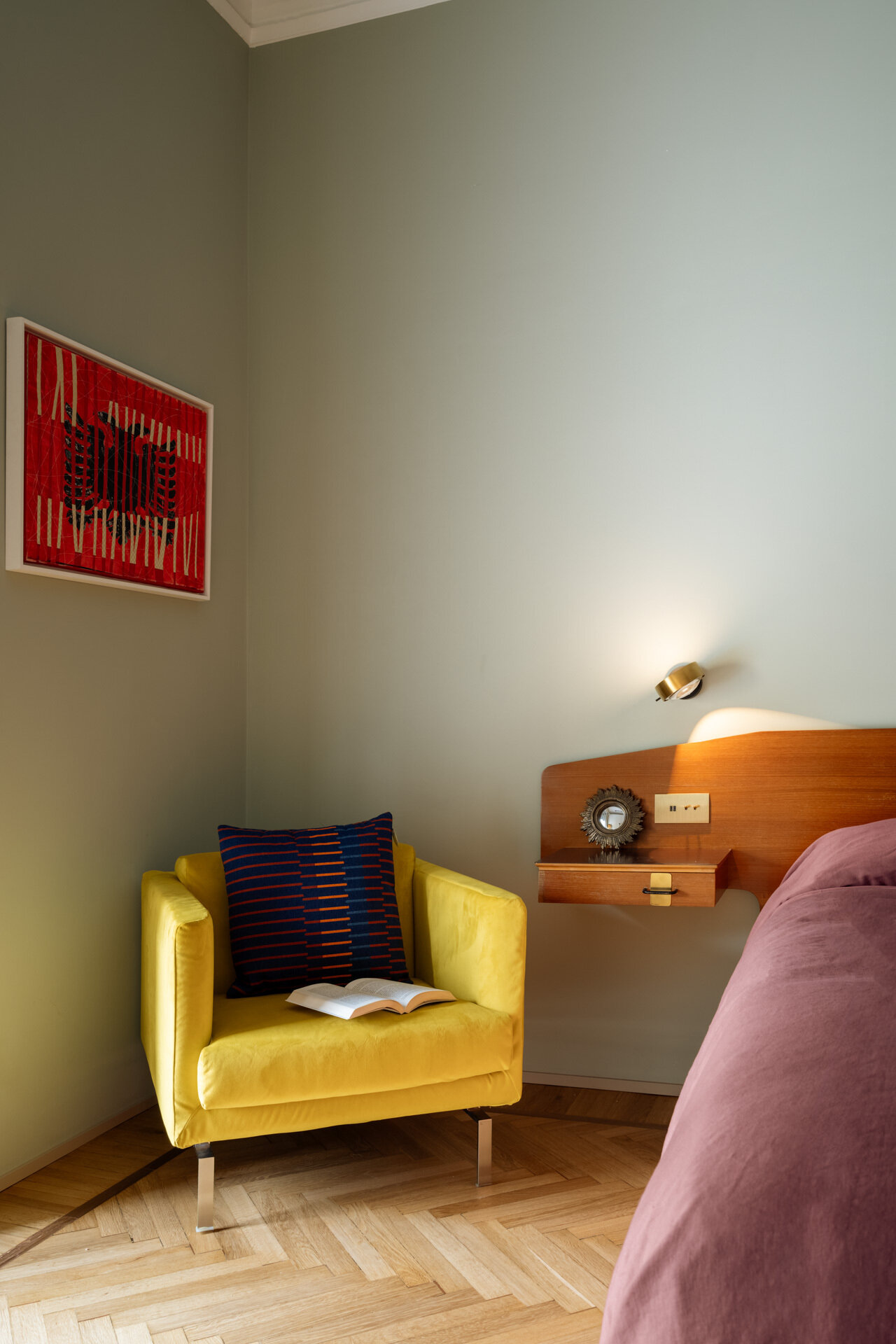 A sage green primary bedroom in Rome combines a vintage teak headboard with brass details, integrated bedside drawers, Occhio reading lights, mauve bedding, a yellow armchair, and original Italian herringbone parquet flooring.