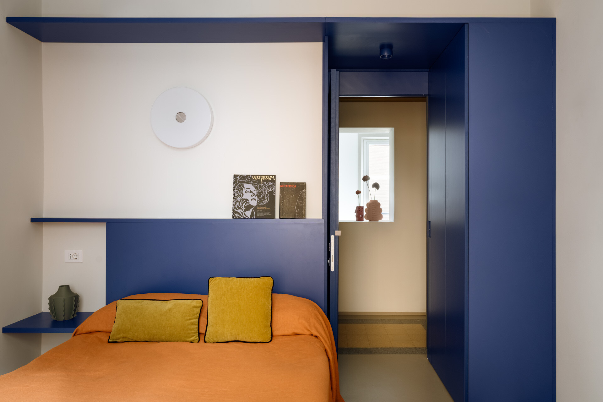 A colorful bedroom in a renovated Rome apartment features a custom peacock-blue headboard, integrated shelves and storage, orange bedding, yellow cushions, a vintage Flos lamp, and original geometric flooring.
