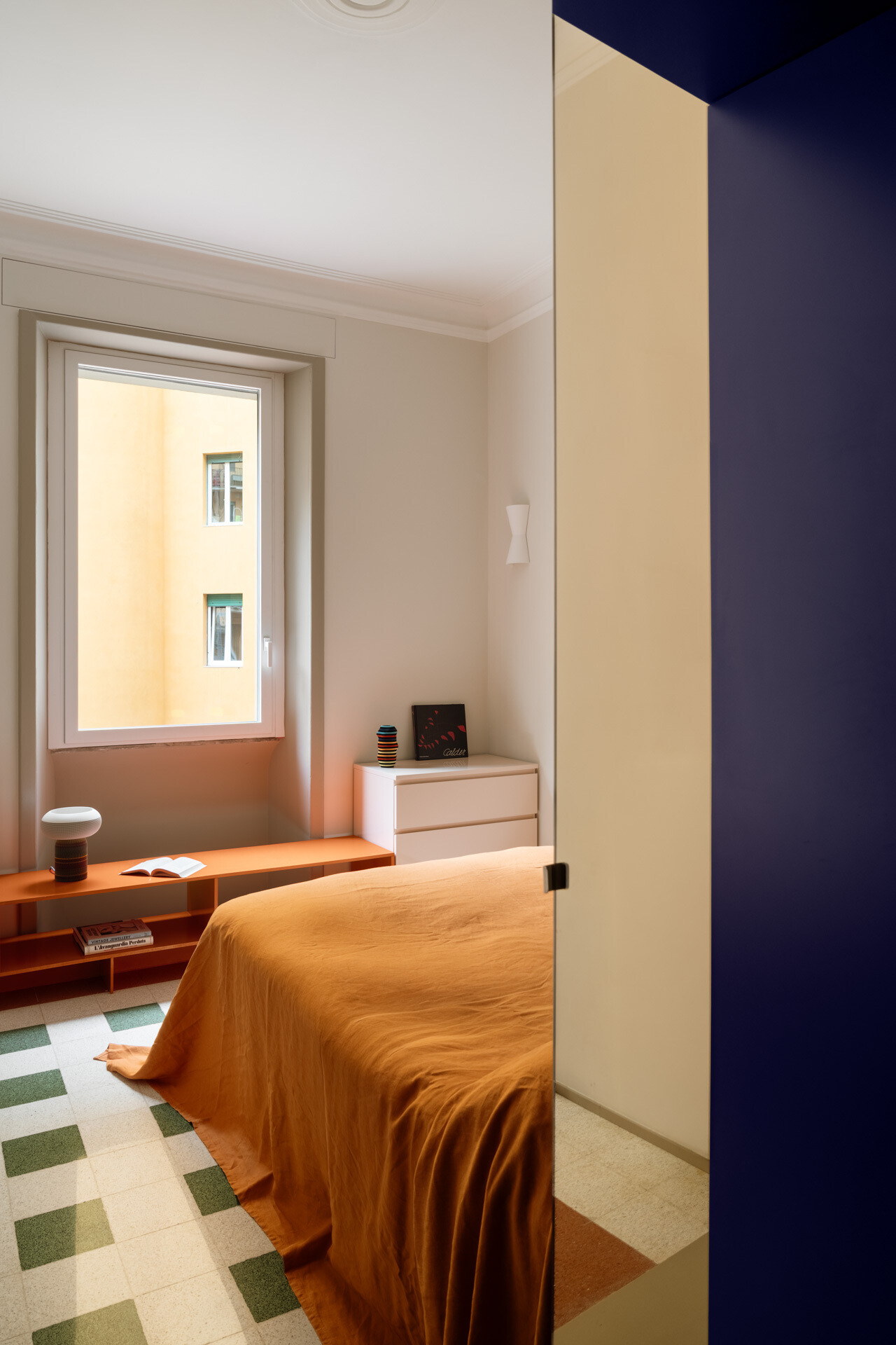 A colorful bedroom in a renovated Rome apartment features a custom peacock-blue headboard, integrated shelves and storage, orange bedding, yellow cushions, a vintage Flos lamp, and original geometric flooring.