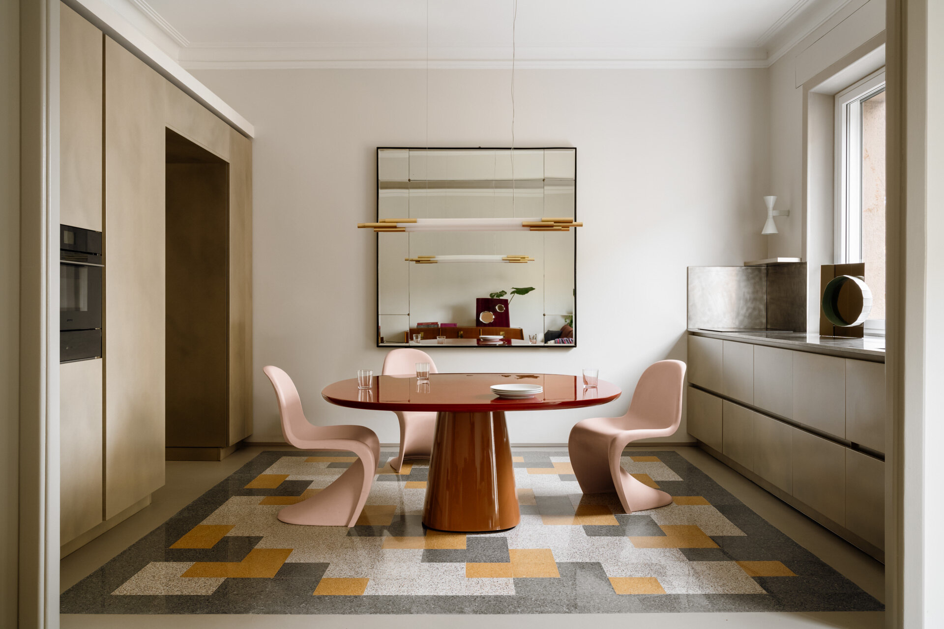 This contemporary kitchen and dining room in Rome features a glossy two-tone B&B Italia dining table, pale pink Vitra Panton Chairs, a gold-lacquered Arrital kitchen, brushed-steel countertops, and restored geometric graniglia flooring.