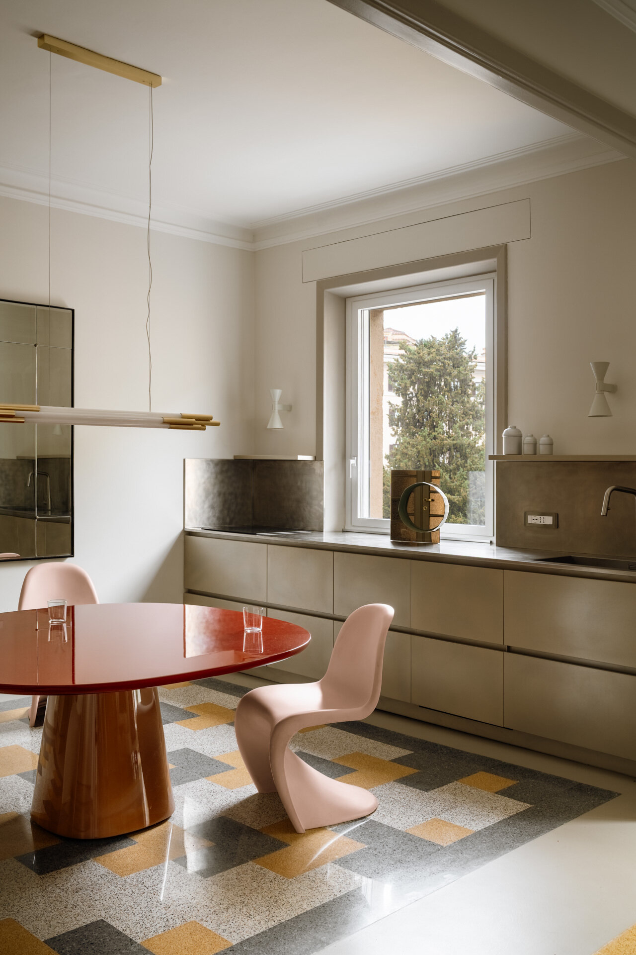 This contemporary kitchen and dining room in Rome features a glossy two-tone B&B Italia dining table, pale pink Vitra Panton Chairs, a gold-lacquered Arrital kitchen, brushed-steel countertops, and restored geometric graniglia flooring.