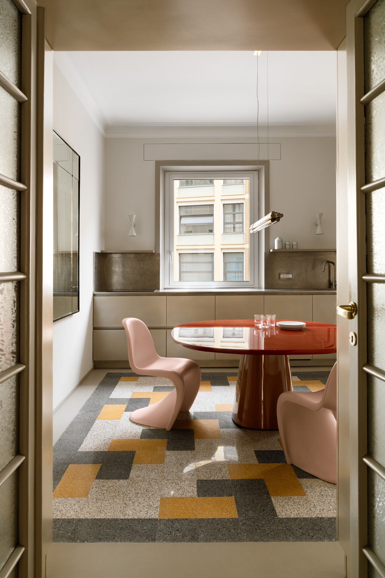 This contemporary kitchen and dining room in Rome features a glossy two-tone B&B Italia dining table, pale pink Vitra Panton Chairs, a gold-lacquered Arrital kitchen, brushed-steel countertops, and restored geometric graniglia flooring.