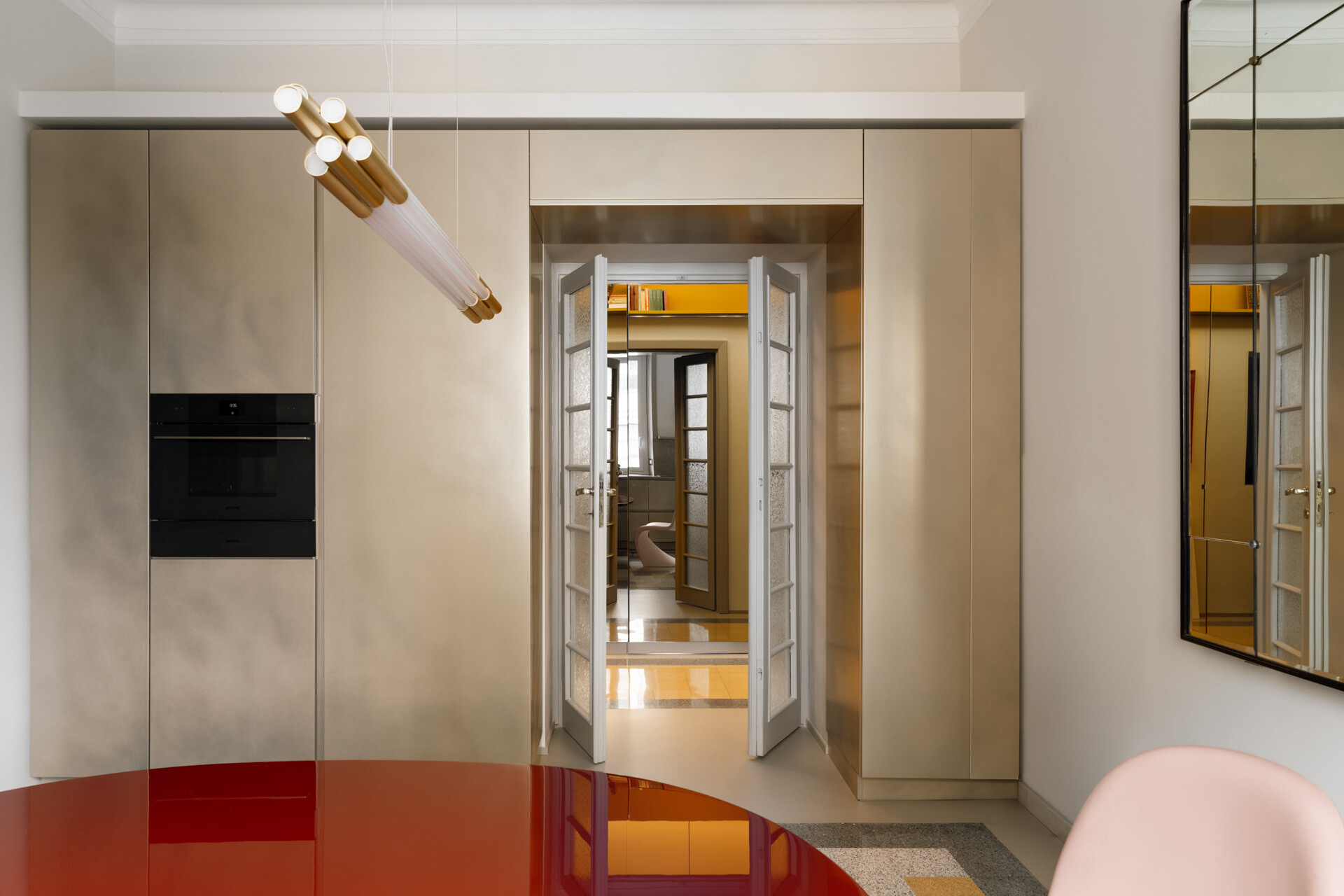 This contemporary kitchen and dining room in Rome features a glossy two-tone B&B Italia dining table, pale pink Vitra Panton Chairs, a gold-lacquered Arrital kitchen, brushed-steel countertops, and restored geometric graniglia flooring.