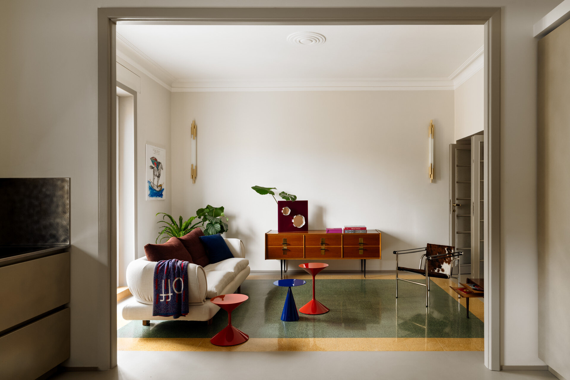 A colorful living room in a renovated 1930s Rome apartment features original green and yellow graniglia flooring, a white Dema sofa, sculptural Zanotta coffee tables, an LC1 armchair by Cassina, and vintage furniture.