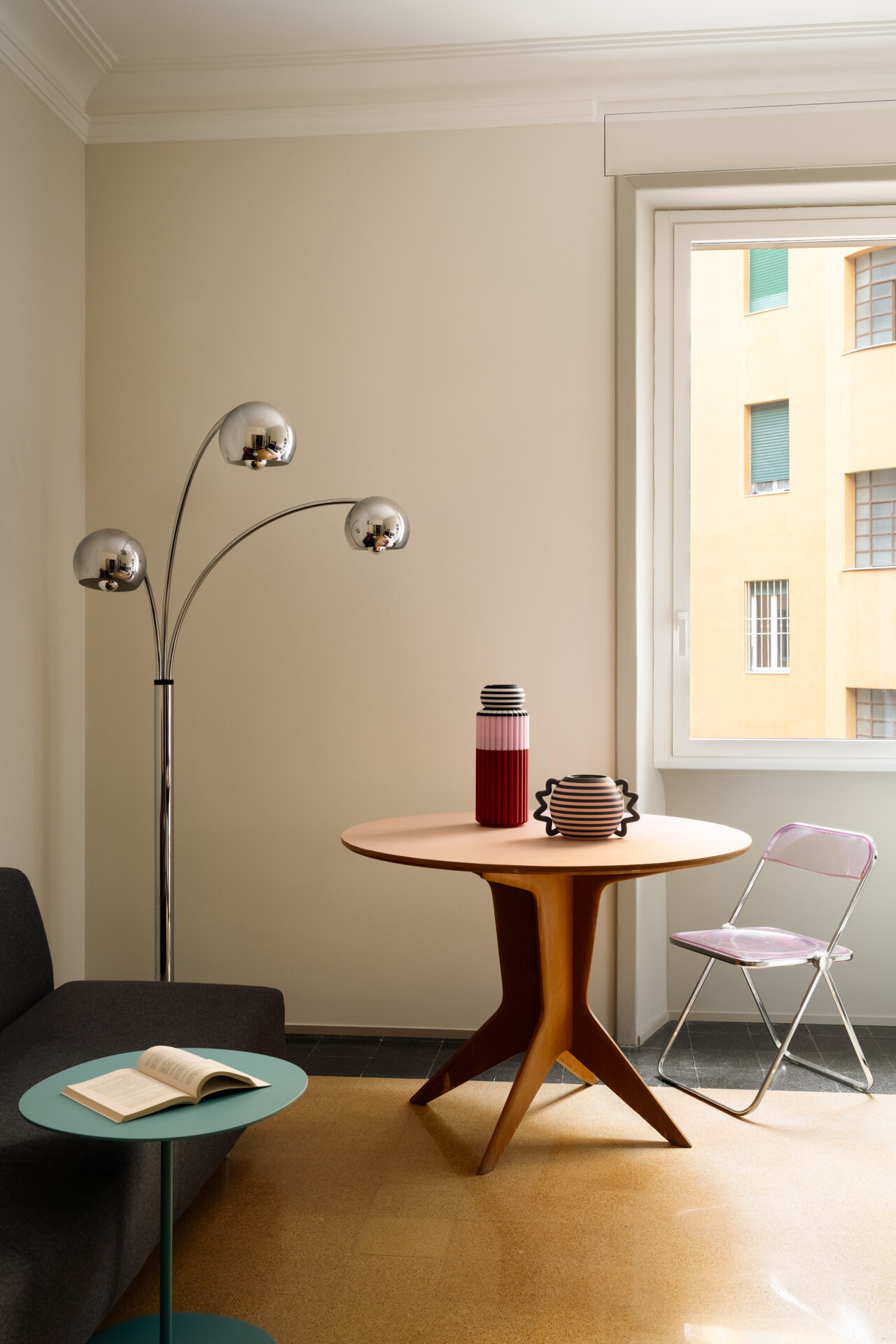 A small sitting room in a renovated Rome apartment combines a round table, transparent pink folding chair, chrome floor lamp, dark sofa, and turquoise side table with original 1930s architectural details.