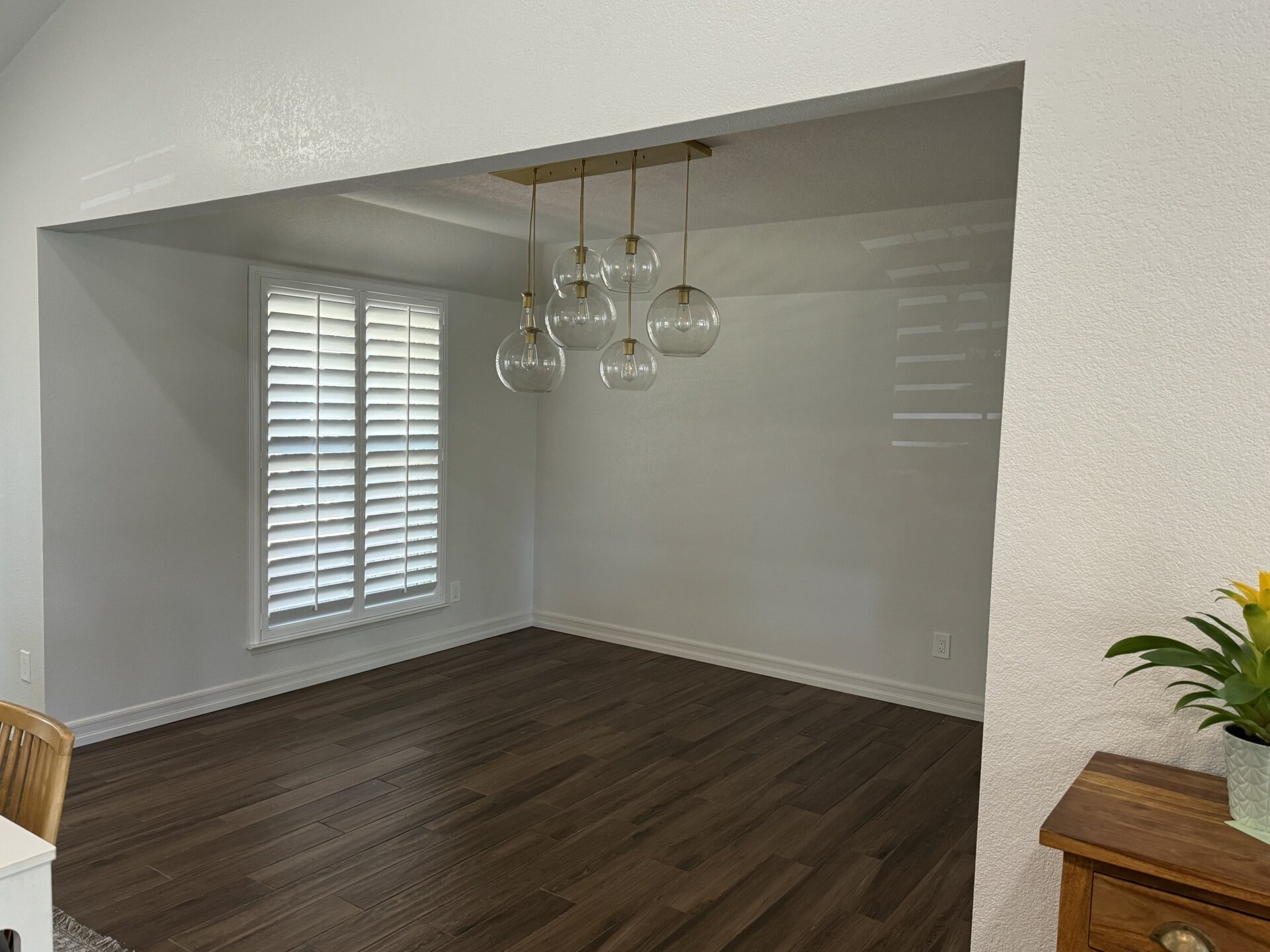 An original dining room in a California ranch house before its conversion into a home office. The open space features white walls, dark plank flooring, window shutters, and a brass pendant light with clear glass globes.