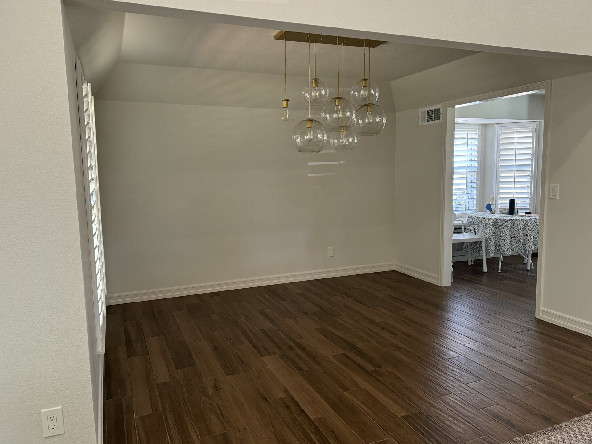 An original dining room in a California ranch house before its conversion into a home office. The open space features white walls, dark plank flooring, window shutters, and a brass pendant light with clear glass globes.