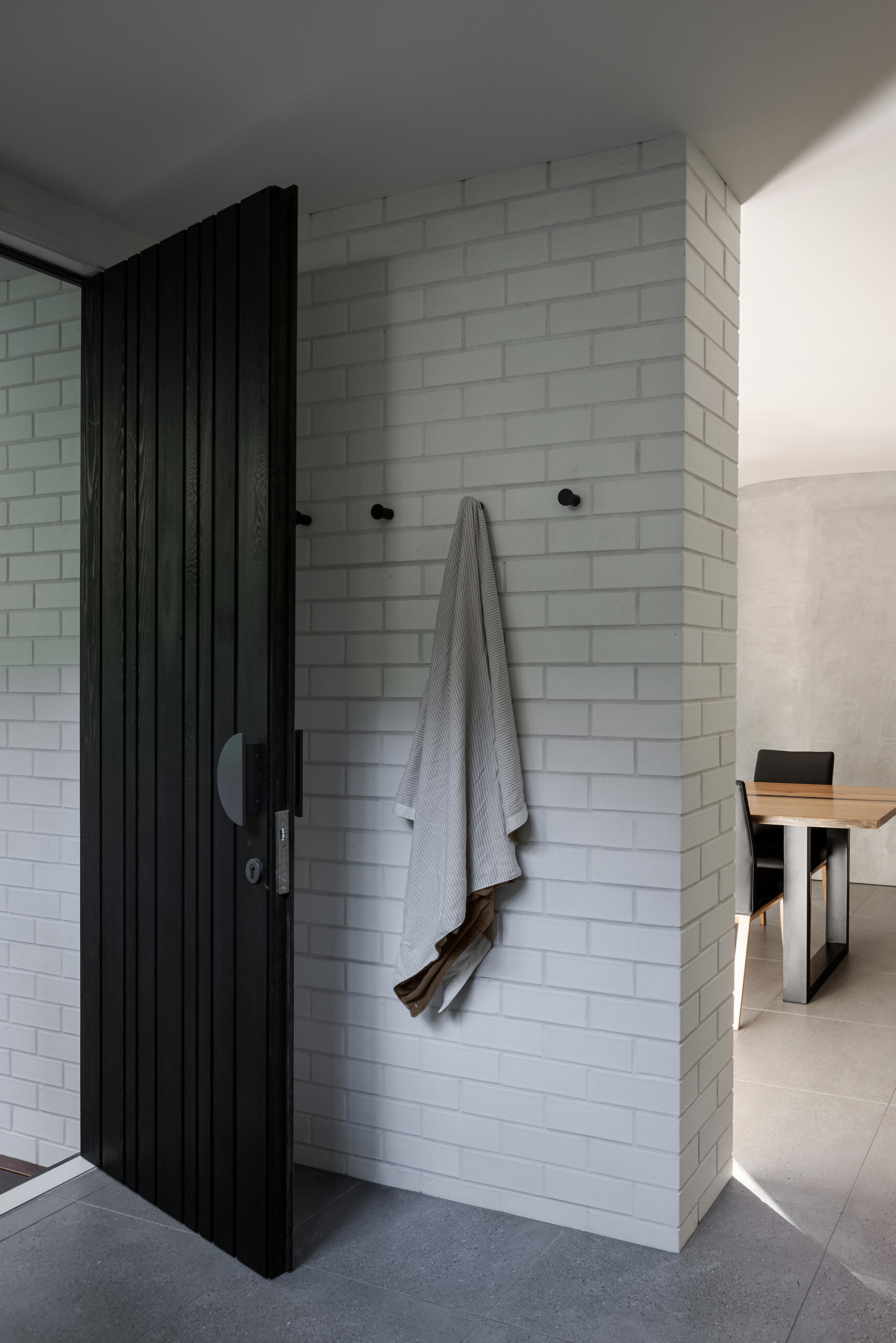 Step inside Raintree in Noosa Heads, Australia, where a black timber front door, white brick walls, minimalist wall hooks, and grey floor tiles create a simple, modern entryway.