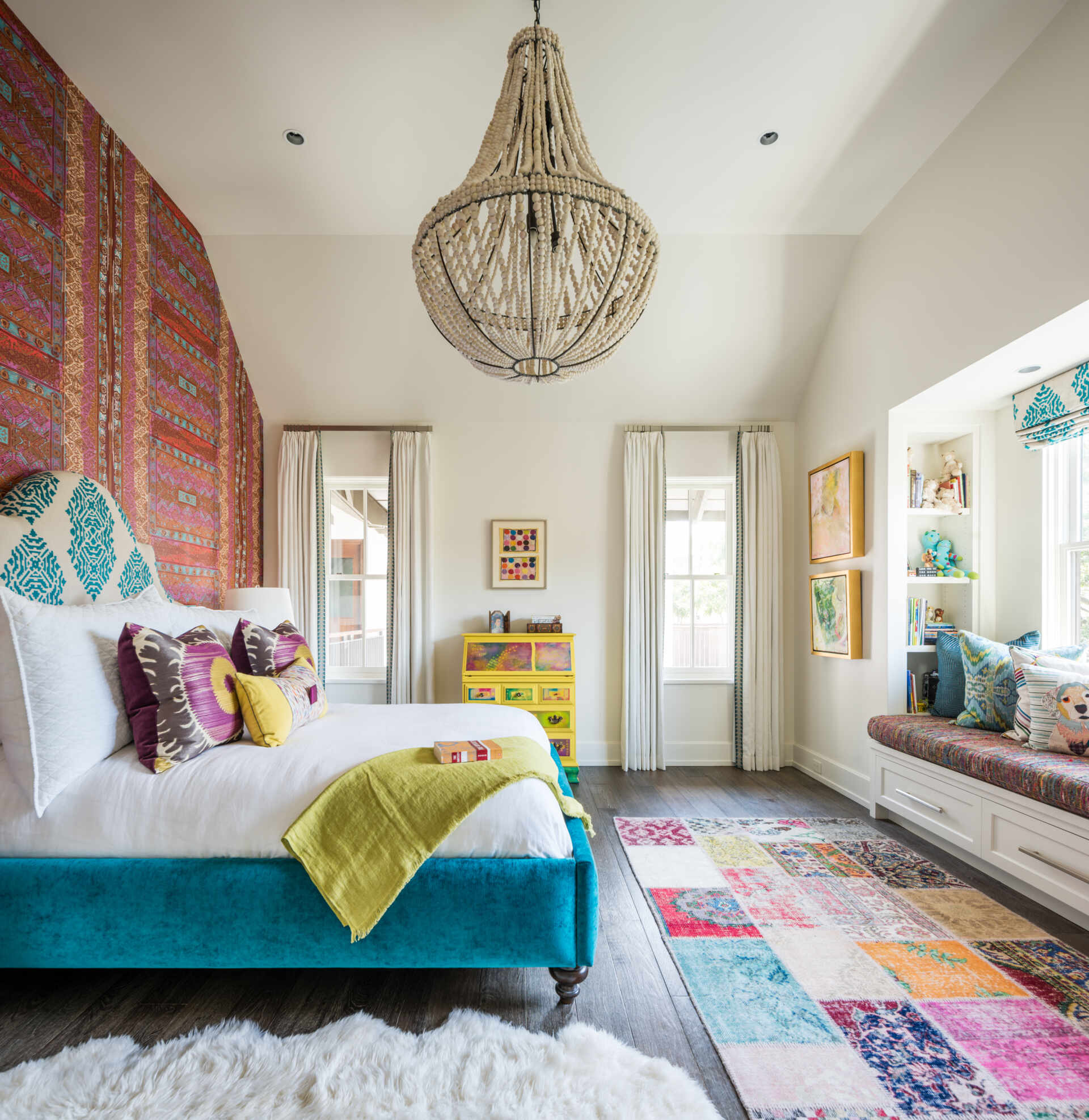 A colorful bedroom with vibrant patterned accent wall, a turquoise upholstered bed, oversized beaded chandelier, multicolored patchwork rug, yellow dresser, and built-in window seat with storage.