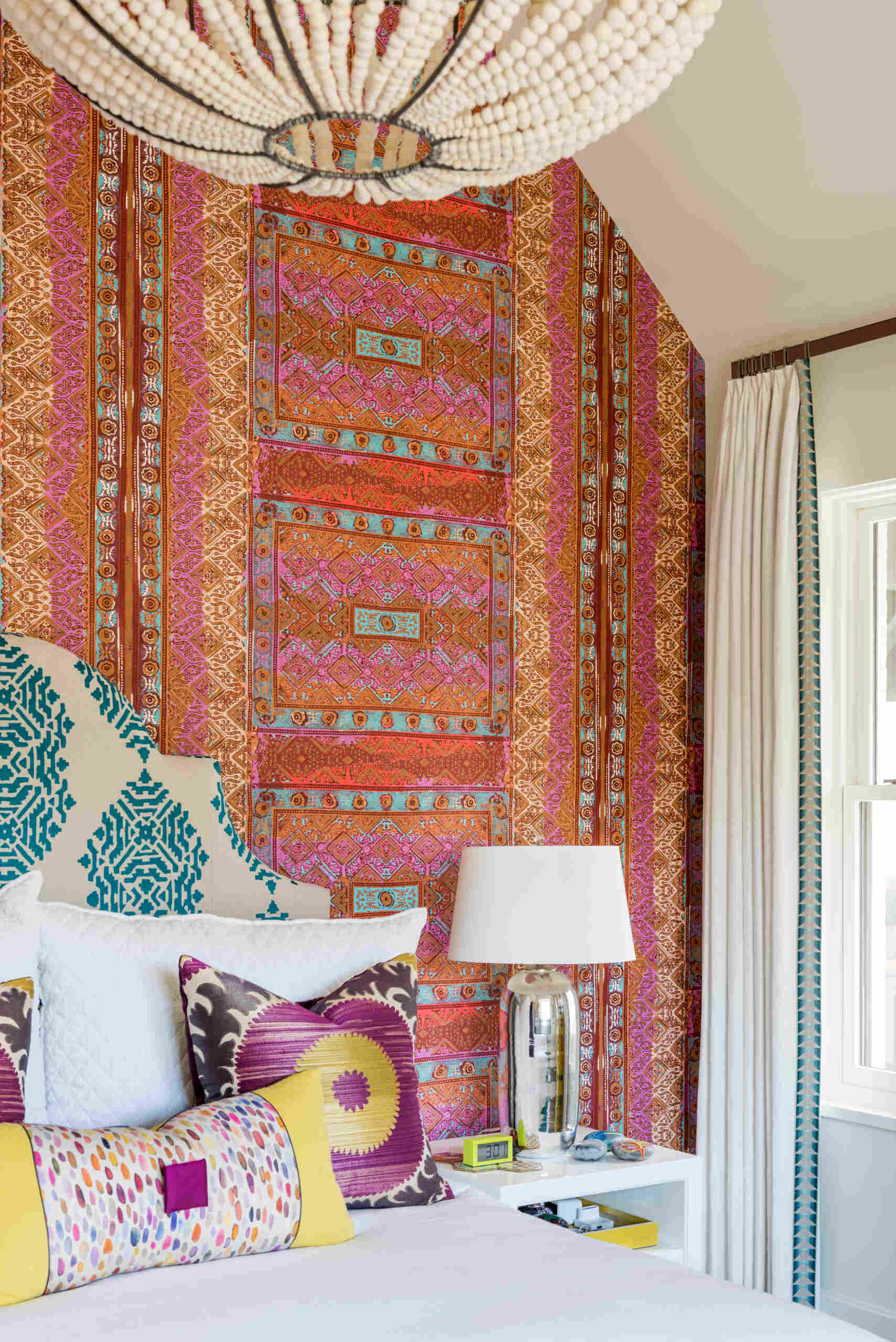 A colorful bedroom with vibrant patterned accent wall, a turquoise upholstered bed, oversized beaded chandelier, multicolored patchwork rug, yellow dresser, and built-in window seat with storage.