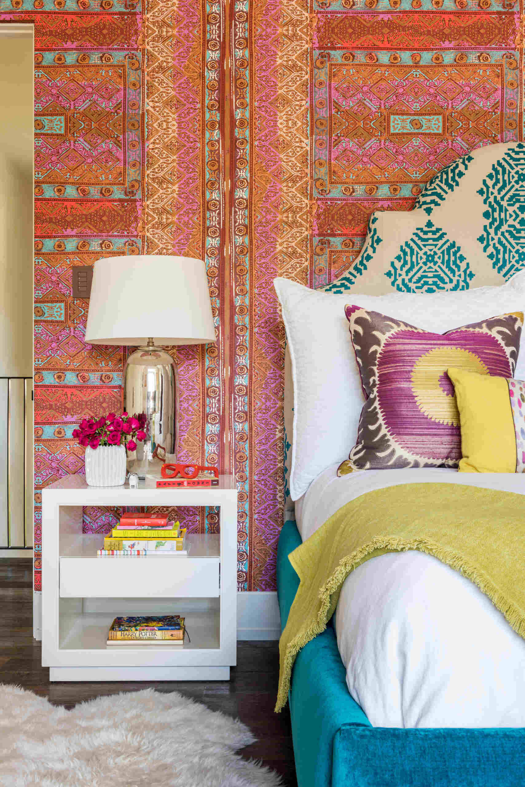 A colorful bedroom with vibrant patterned accent wall, a turquoise upholstered bed, oversized beaded chandelier, multicolored patchwork rug, yellow dresser, and built-in window seat with storage.