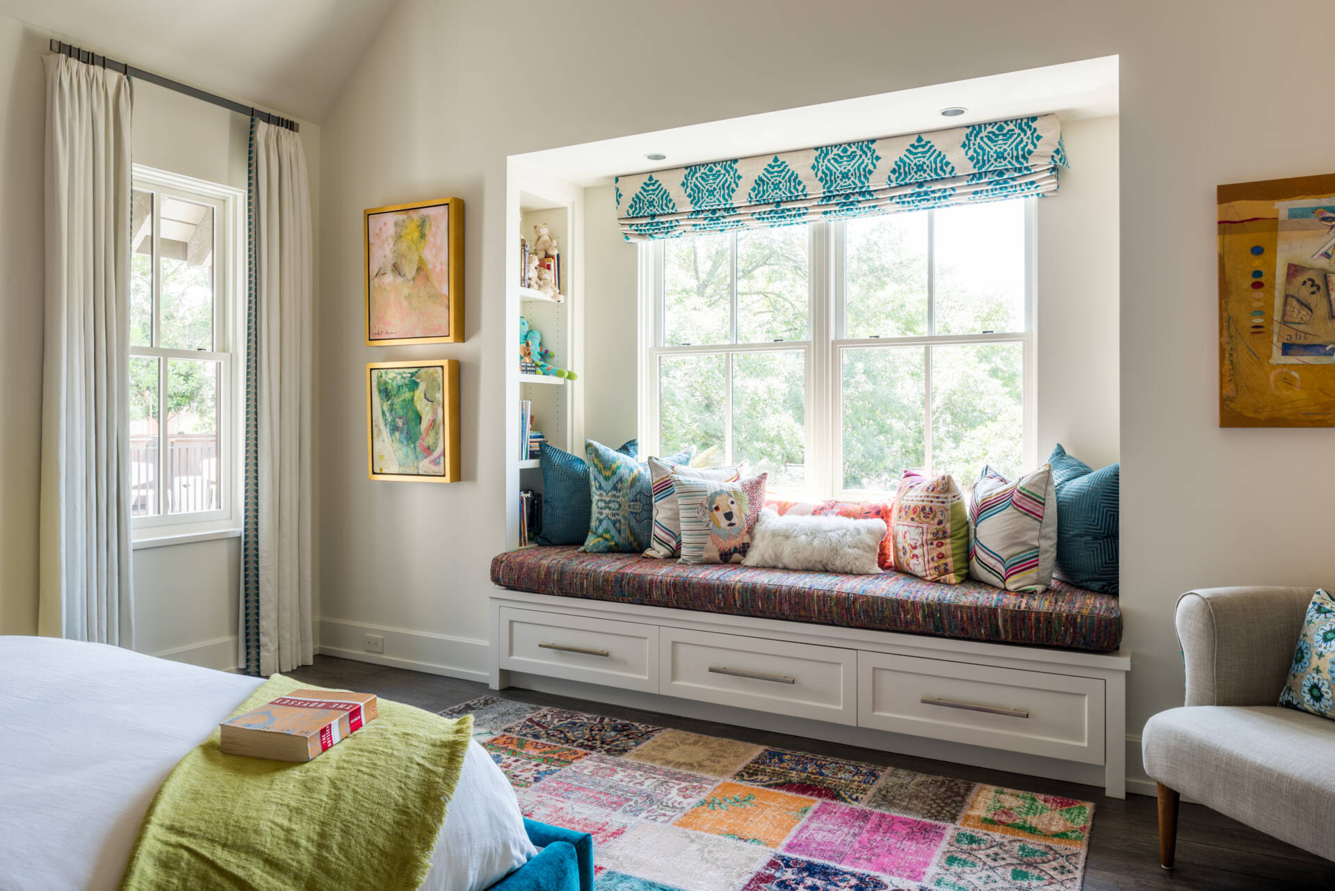 A colorful bedroom with vibrant patterned accent wall, a turquoise upholstered bed, oversized beaded chandelier, multicolored patchwork rug, yellow dresser, and built-in window seat with storage.