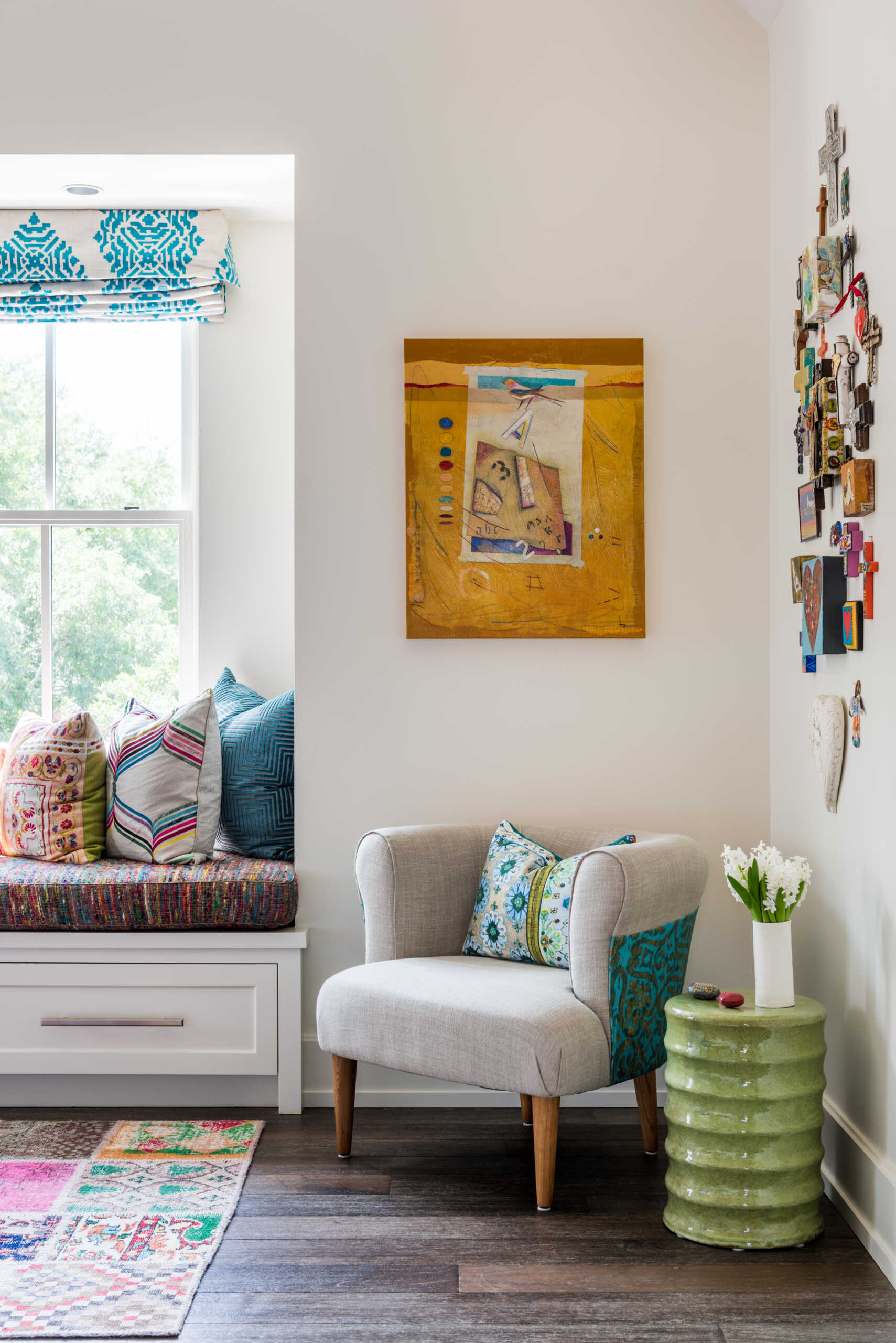 A colorful bedroom with vibrant patterned accent wall, a turquoise upholstered bed, oversized beaded chandelier, multicolored patchwork rug, yellow dresser, and built-in window seat with storage.