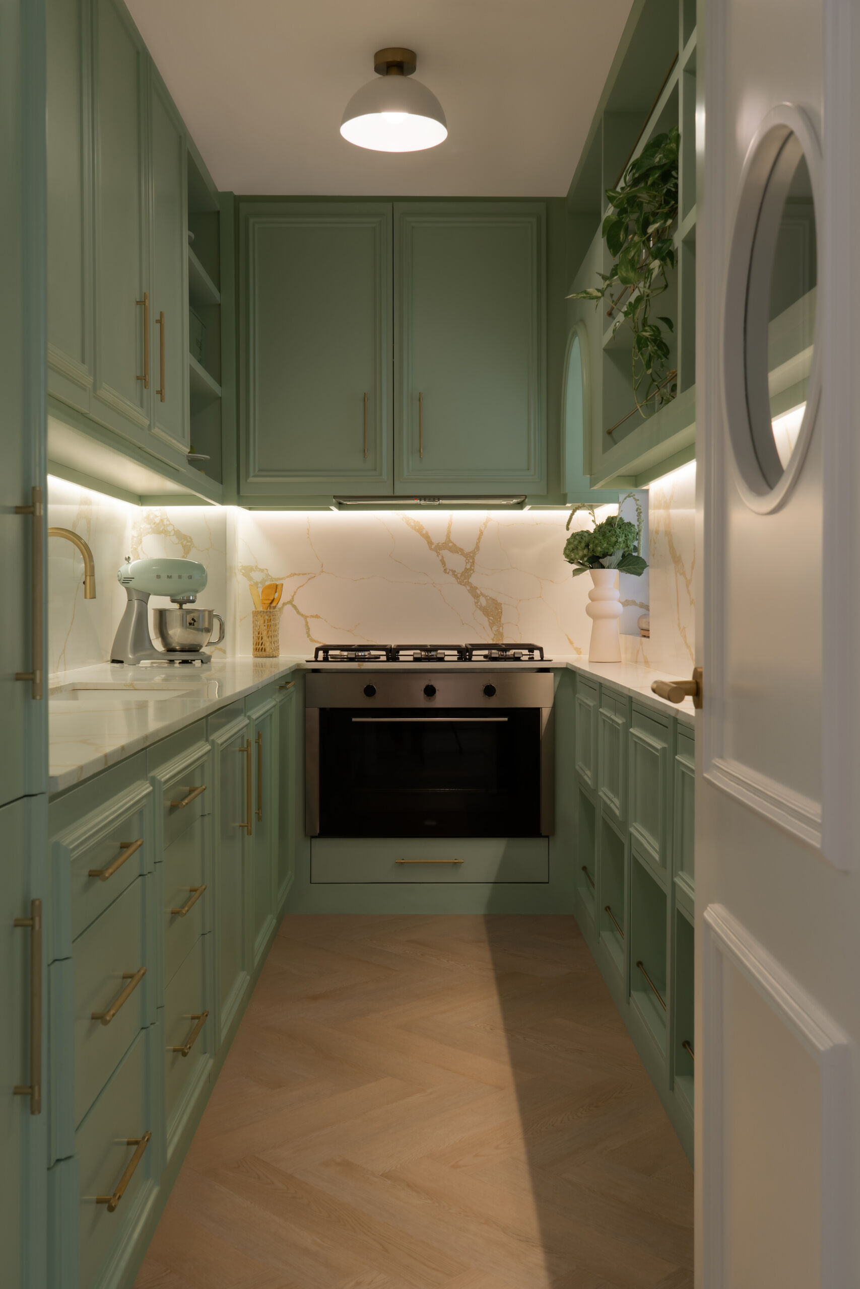 A sage-green butler's pantry adds a beautiful surprise to The White Arcade in Dubai. Designed by D’Ora Tokai Designs, this compact kitchen features green panelled cabinetry, white stone countertops, brass handles, open shelves, and a charming round window in the door.