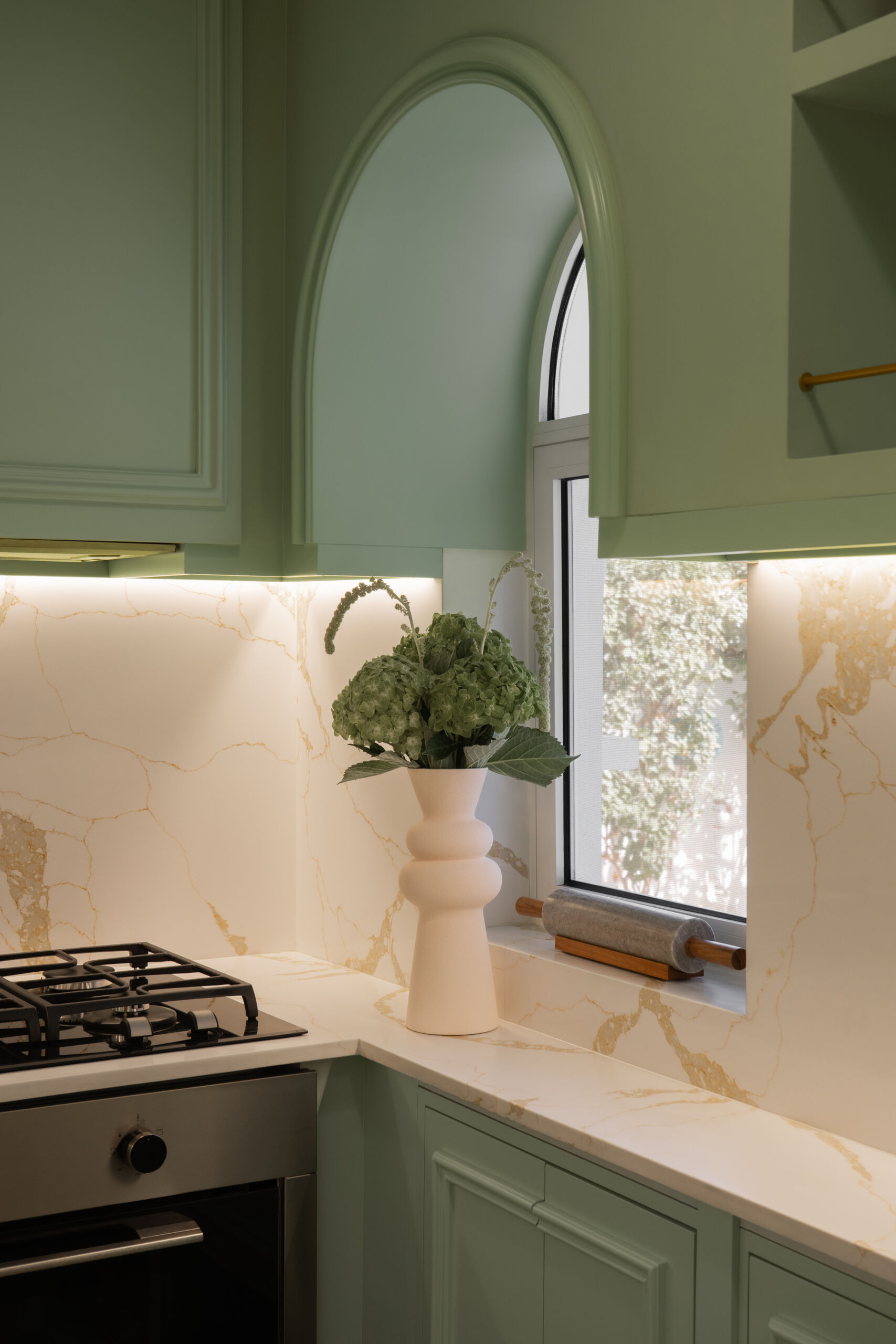 A sage-green butler's pantry adds a beautiful surprise to The White Arcade in Dubai. Designed by D’Ora Tokai Designs, this compact kitchen features green panelled cabinetry, white stone countertops, brass handles, open shelves, and a charming round window in the door.