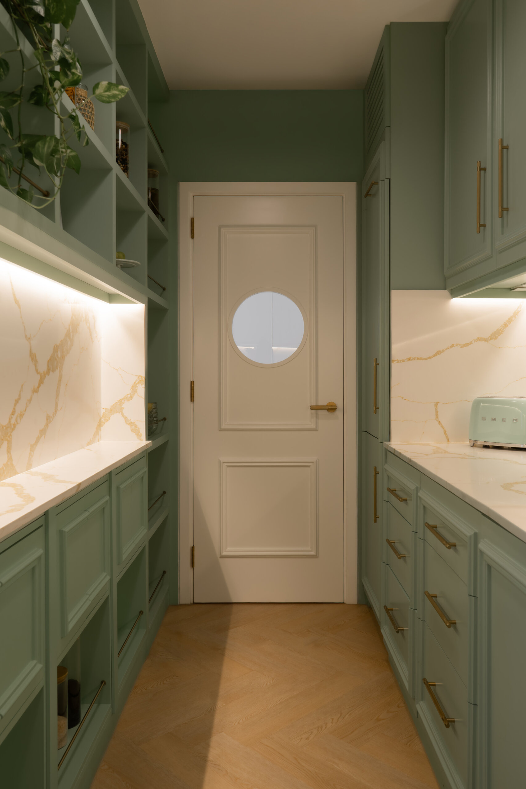 A sage-green butler's pantry adds a beautiful surprise to The White Arcade in Dubai. Designed by D’Ora Tokai Designs, this compact kitchen features green panelled cabinetry, white stone countertops, brass handles, open shelves, and a charming round window in the door.
