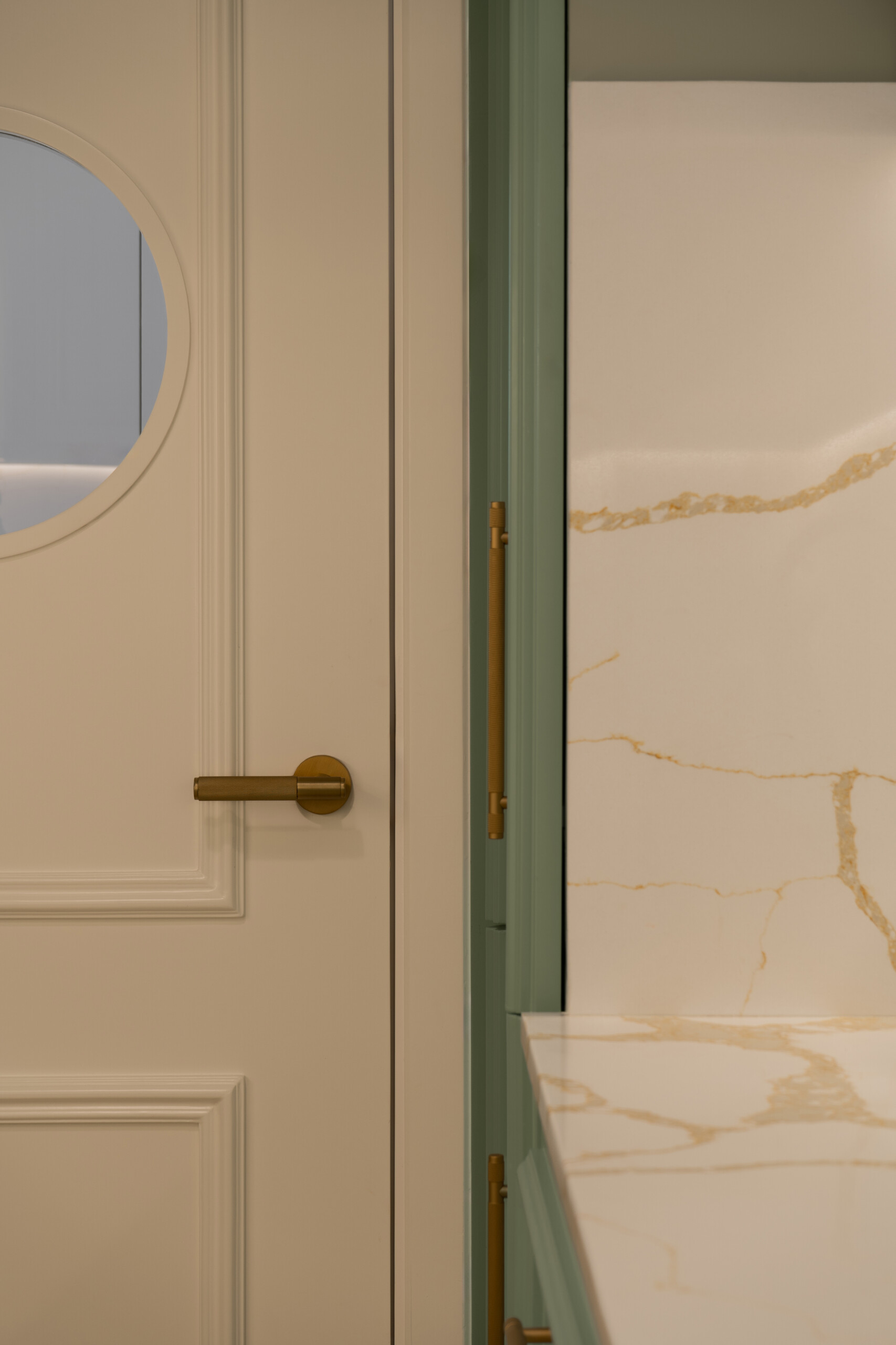 A sage-green butler's pantry adds a beautiful surprise to The White Arcade in Dubai. Designed by D’Ora Tokai Designs, this compact kitchen features green panelled cabinetry, white stone countertops, brass handles, open shelves, and a charming round window in the door.