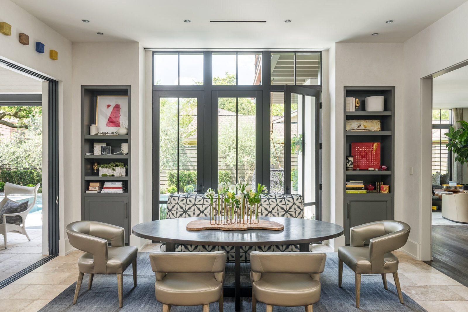 A casual dining room with a dark oval table, upholstered chairs, patterned banquette, built-in gray bookshelves, and tall glass doors overlooking the garden.