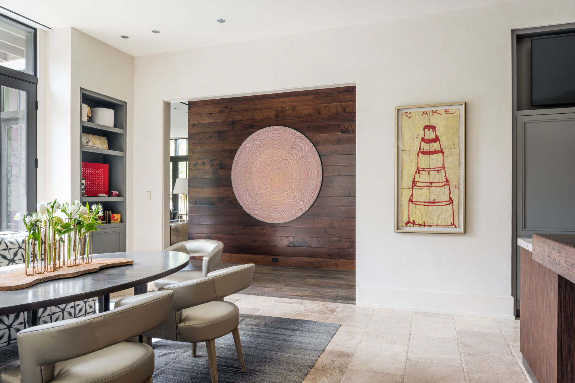 A casual dining room with a dark oval table, upholstered chairs, patterned banquette, built-in gray bookshelves, and tall glass doors overlooking the garden.