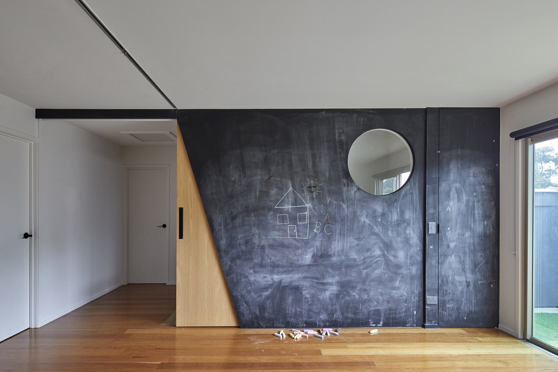 An angled timber sliding door opens to reveal a passage beside a large black chalkboard wall. The design combines a playful drawing surface with a flexible interior doorway.