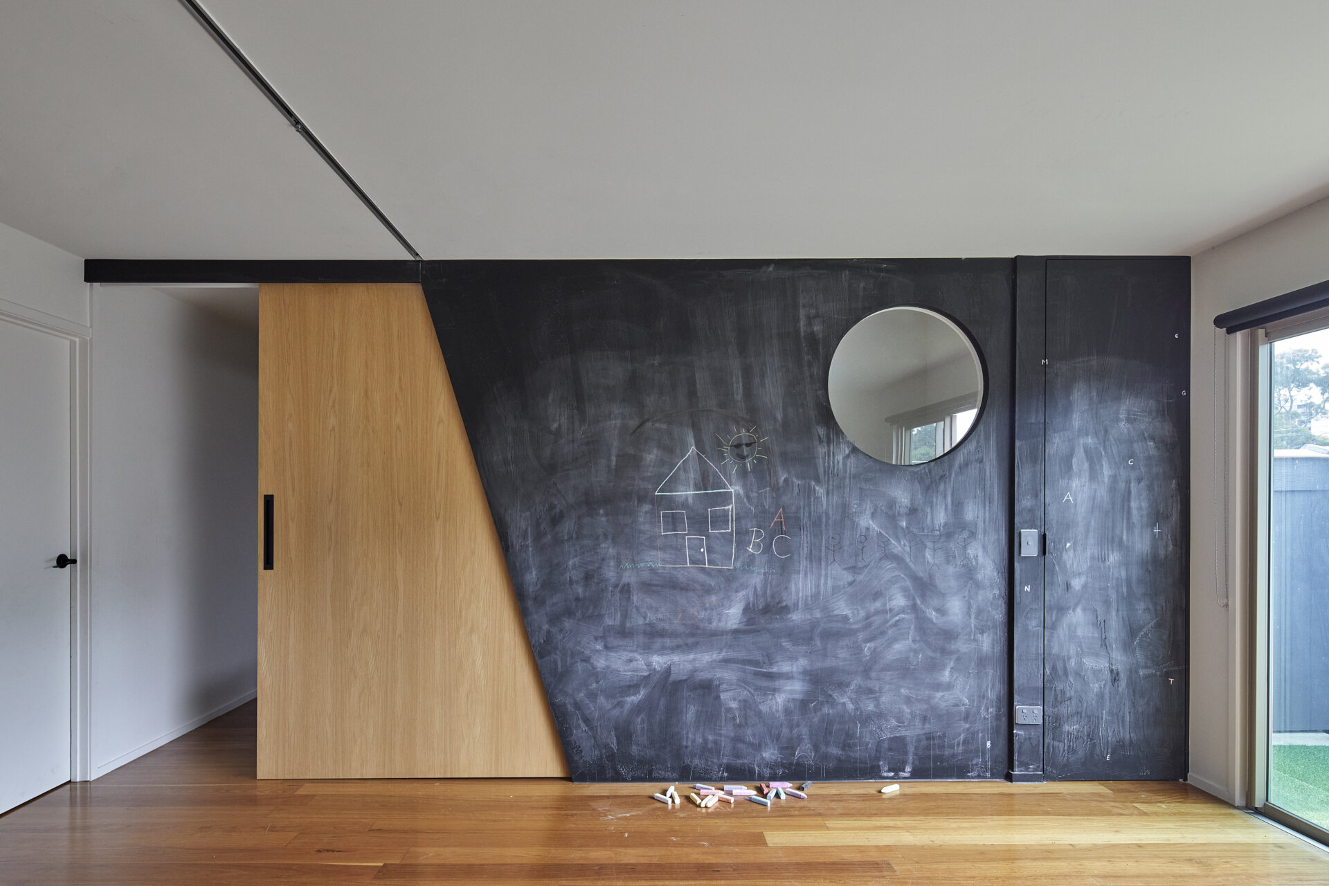 A circular opening provides a view through the chalkboard wall, while a timber panel with an angled edge slides along a ceiling-mounted track to open or close the adjoining passage.