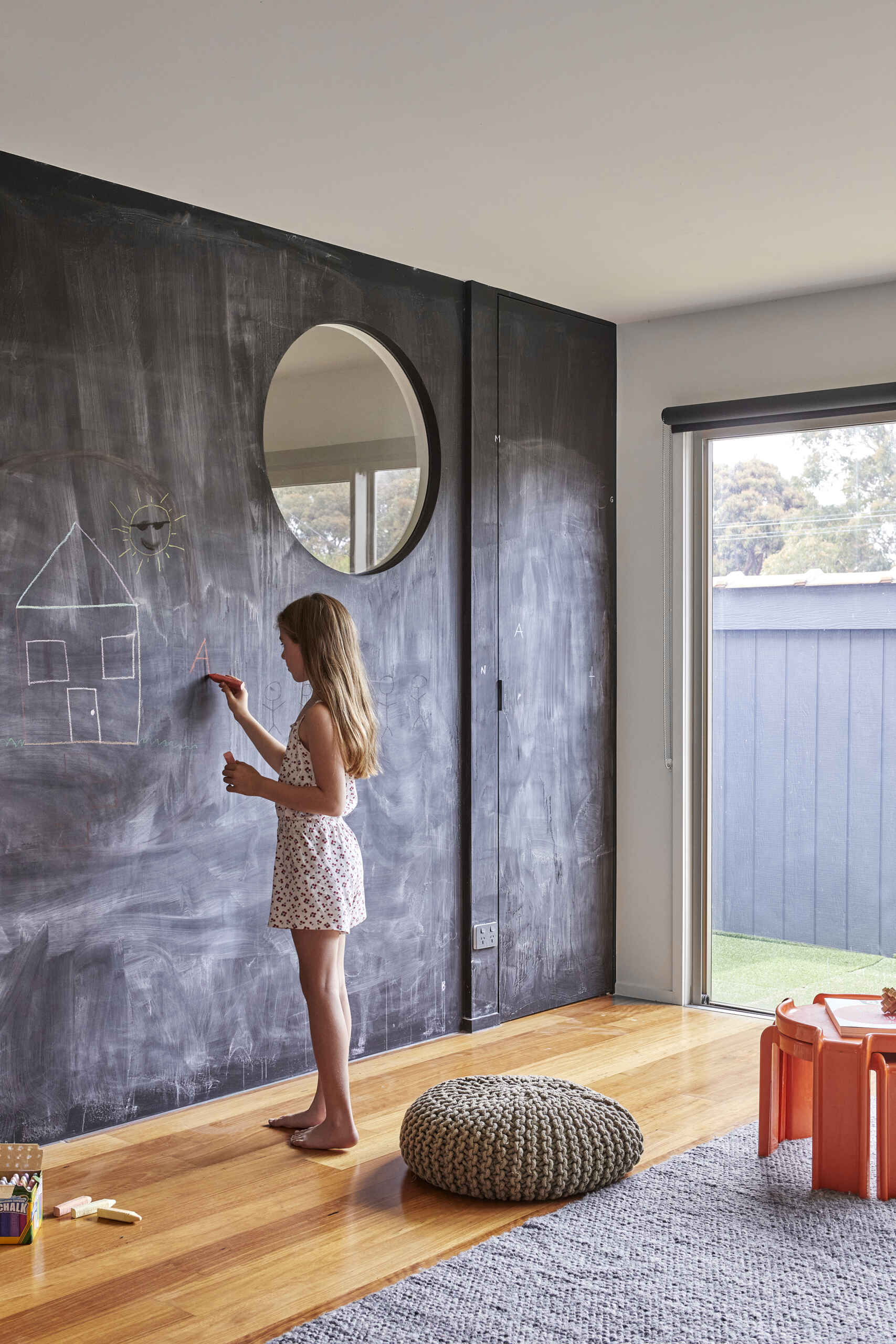A child draws on a floor-to-ceiling chalkboard wall featuring a large circular mirror. The modern playroom also includes timber flooring, a knitted pouf, and a small orange table.