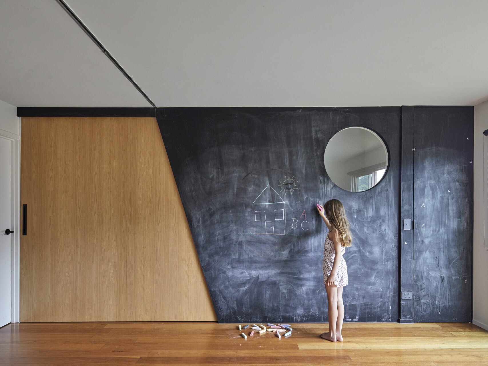 A large black chalkboard wall with children's drawings, a circular mirror, and an angled timber sliding door creates a playful feature in this renovated family home.