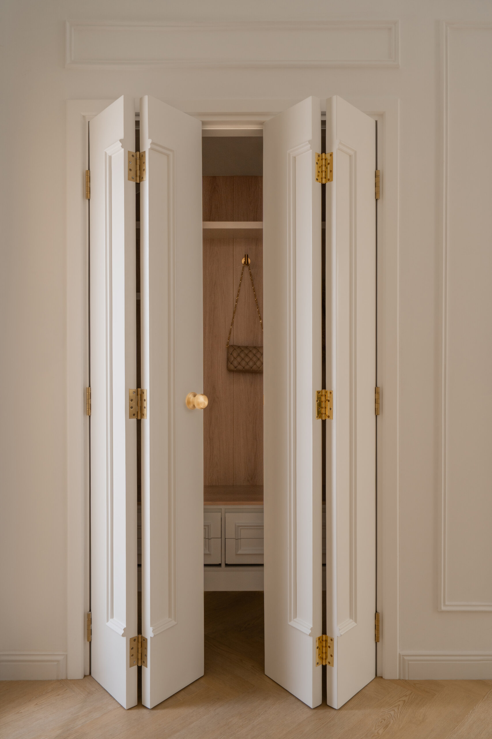 Hidden behind elegant bifold doors, this coat room at The White Arcade in Dubai features oak-lined walls, built-in bench seating, concealed drawers, brass hooks, and practical shelving. A stylish small-space storage idea by D’Ora Tokai Designs.
