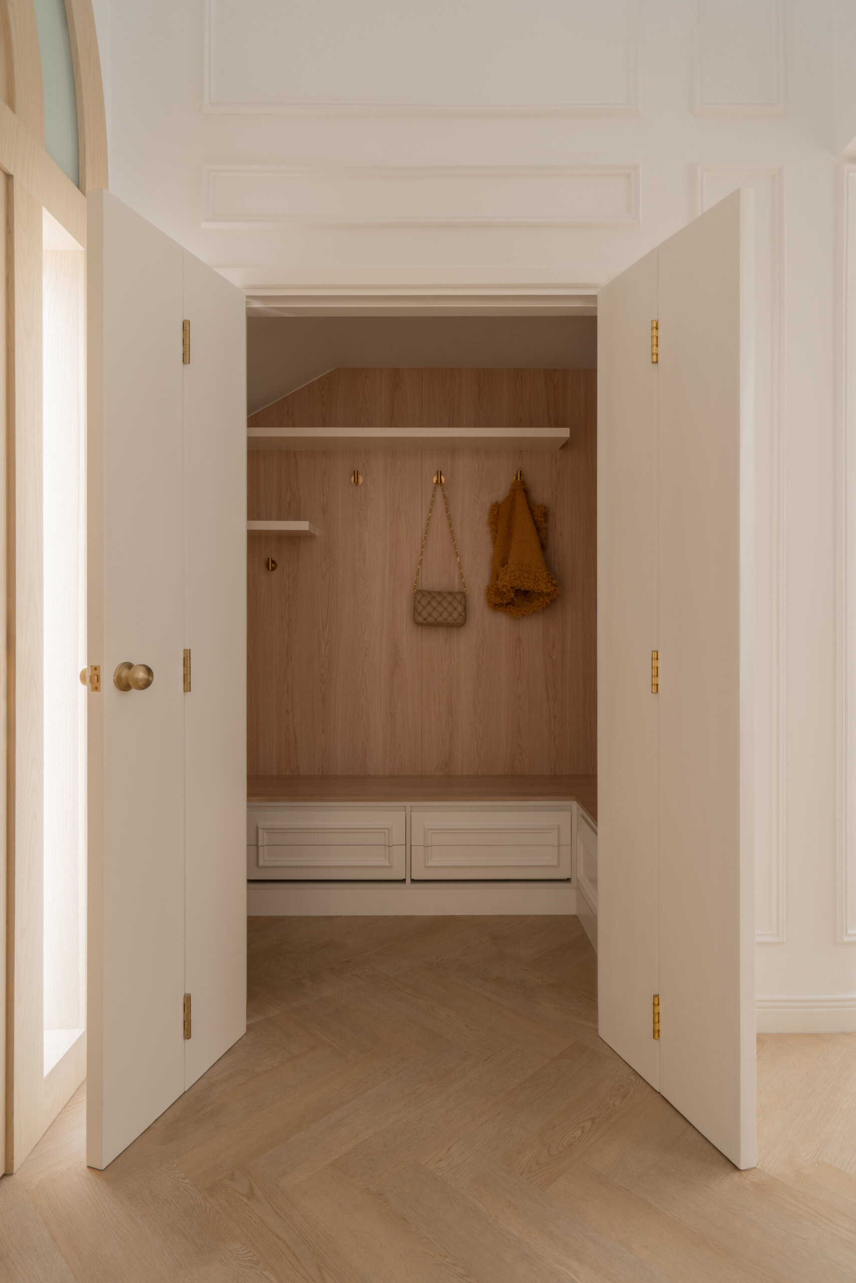 Hidden behind elegant bifold doors, this coat room at The White Arcade in Dubai features oak-lined walls, built-in bench seating, concealed drawers, brass hooks, and practical shelving. A stylish small-space storage idea by D’Ora Tokai Designs.