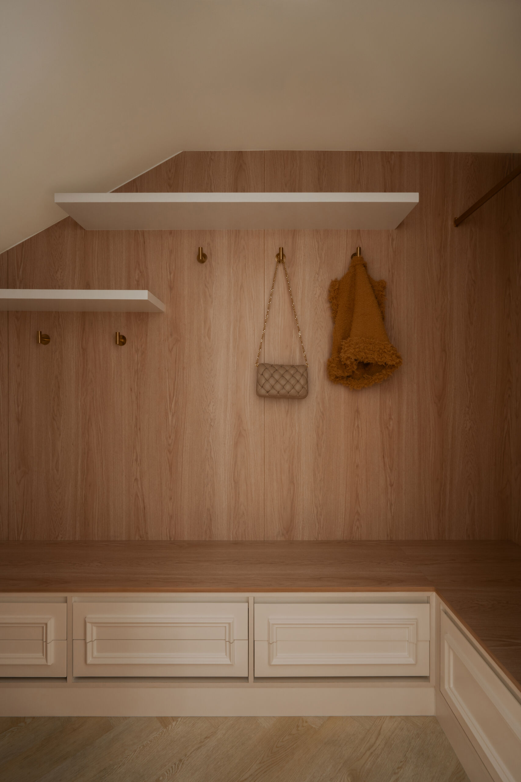 Hidden behind elegant bifold doors, this coat room at The White Arcade in Dubai features oak-lined walls, built-in bench seating, concealed drawers, brass hooks, and practical shelving. A stylish small-space storage idea by D’Ora Tokai Designs.