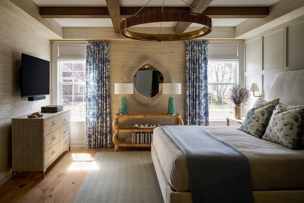 A coastal-inspired primary bedroom with blue and white floral curtains, textured neutral wallpaper, exposed ceiling beams, an upholstered bed, a large round mirror, and turquoise table lamps.
