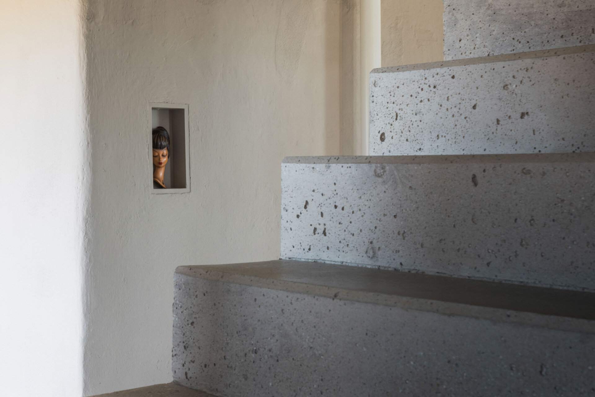 A sculptural concrete staircase in an Austrian farmhouse renovation features angular steps, a pale pink curved handrail, and a custom storage bench with upholstered seating.