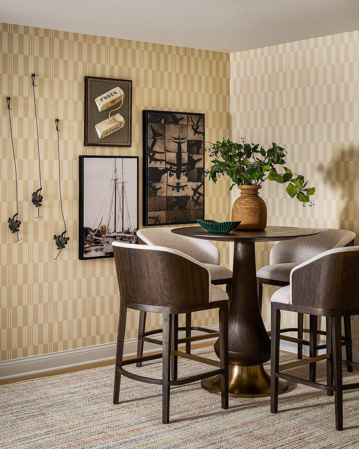 A contemporary bar area with cream geometric wallpaper, a round dark wood pedestal table, upholstered wood barstools, and a gallery wall featuring framed aviation artwork and small sculptural pieces.