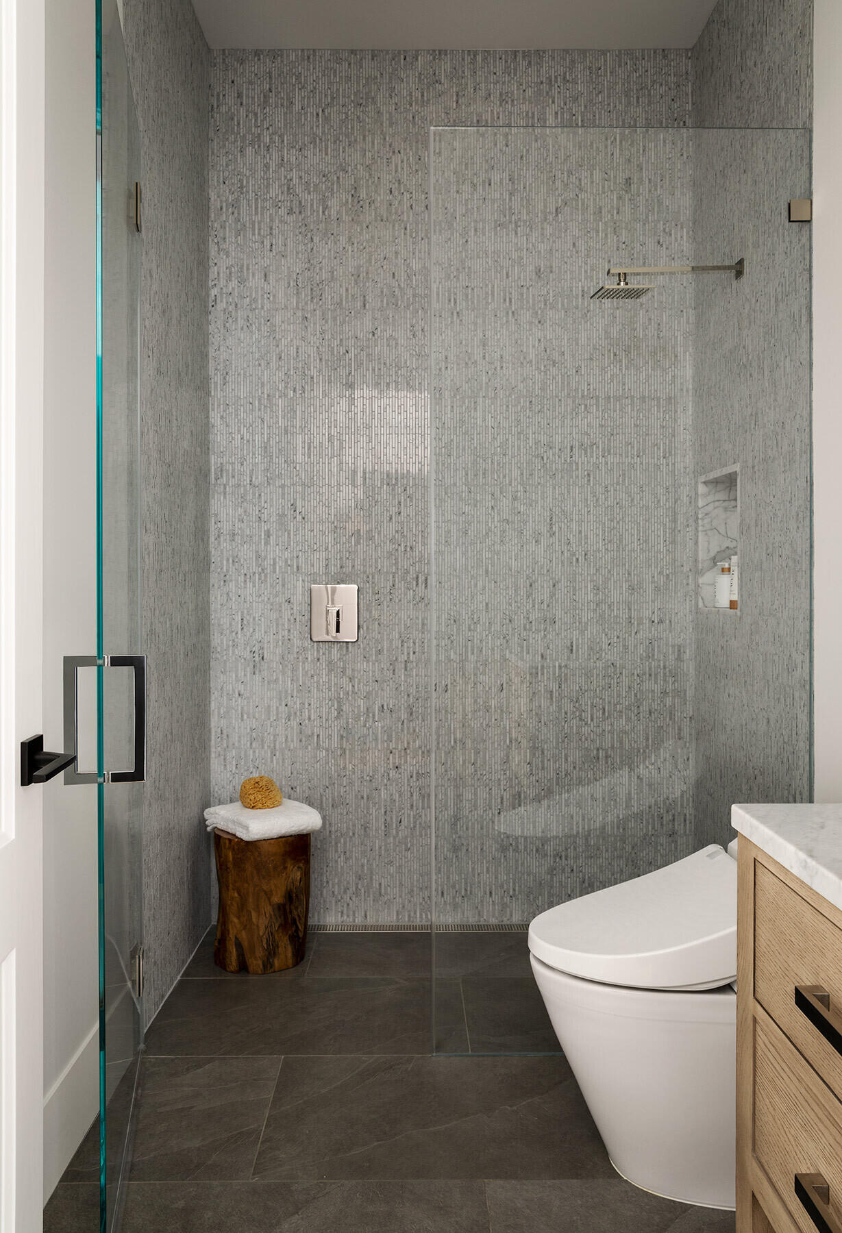 This compact bathroom in the Atherton Residence pairs grey mosaic shower walls, dark stone flooring, frameless glass, a marble-lined niche and light timber cabinetry for a clean, modern finish.