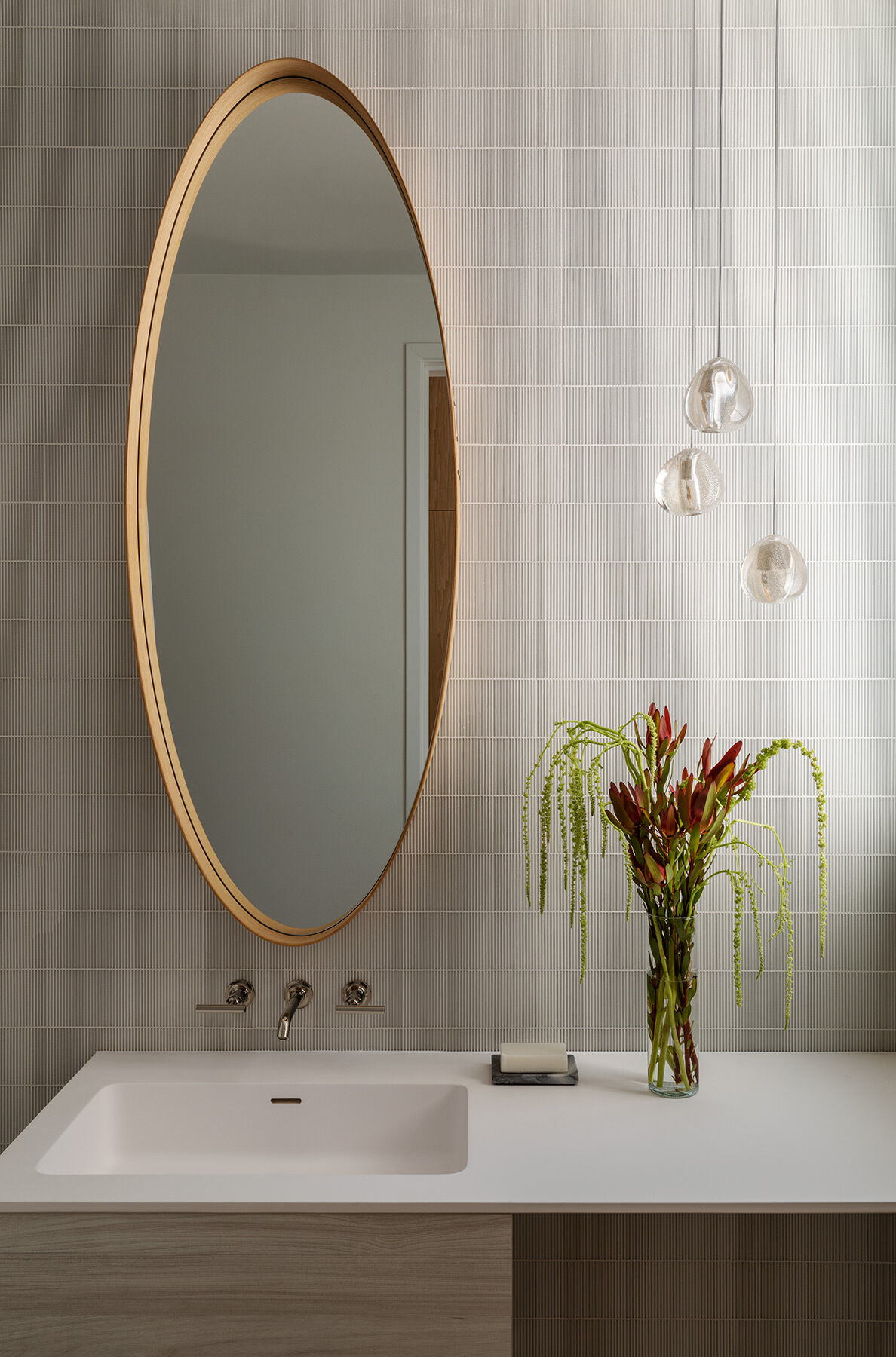 This Atherton Residence powder room pairs fine grey wall tile with an oval timber-framed mirror, wall-mounted faucet, pale vanity and delicate glass pendant lights for a warm modern look.