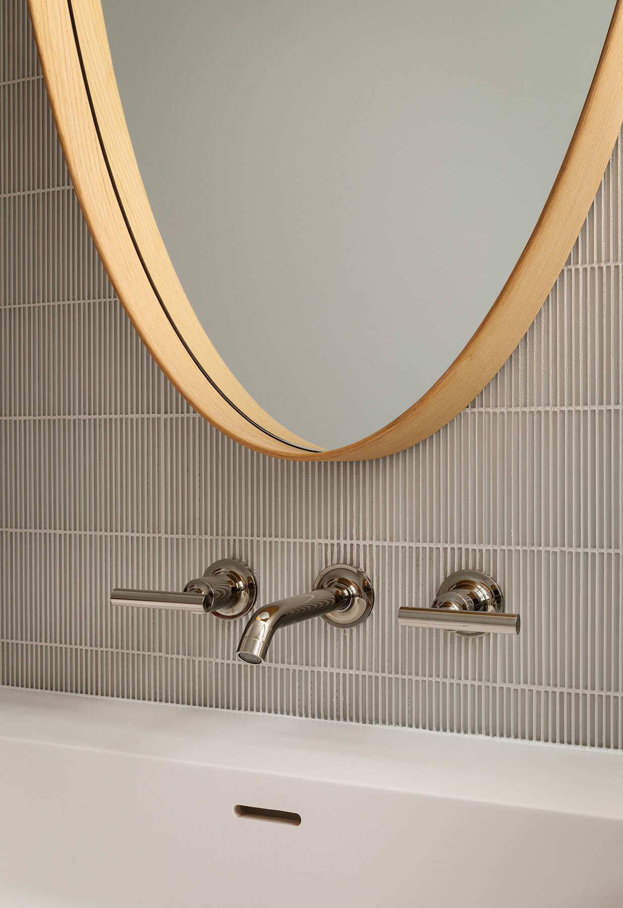This Atherton Residence powder room pairs fine grey wall tile with an oval timber-framed mirror, wall-mounted faucet, pale vanity and delicate glass pendant lights for a warm modern look.