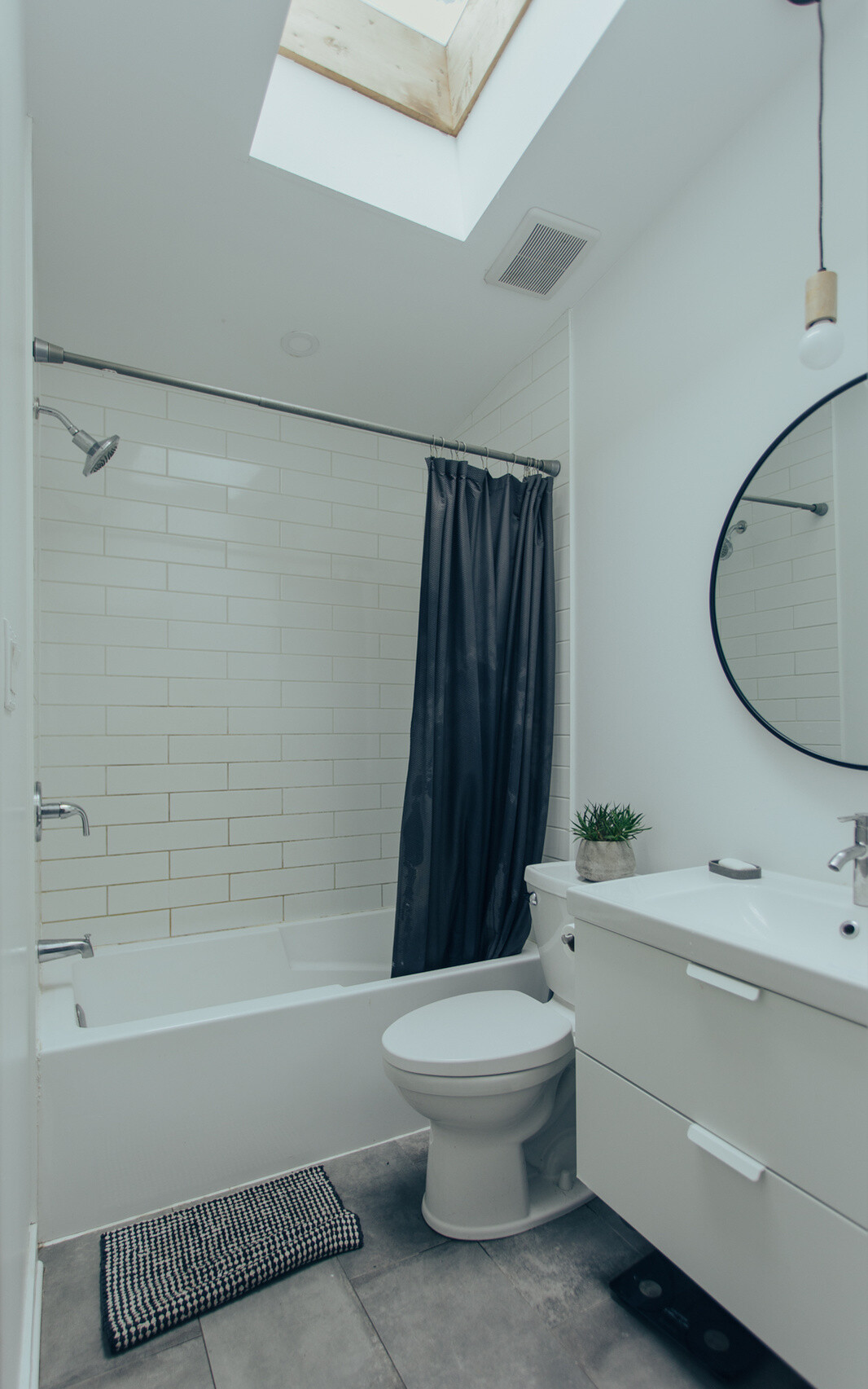 This bathroom has white subway tiles surrounding the bathtub and shower, a compact vanity, and a round black-framed mirror. A skylight positioned above the bathing area introduces daylight from overhead, helping brighten the narrow space. Grey floor tiles and a dark shower curtain provide contrast against the predominantly white finishes.